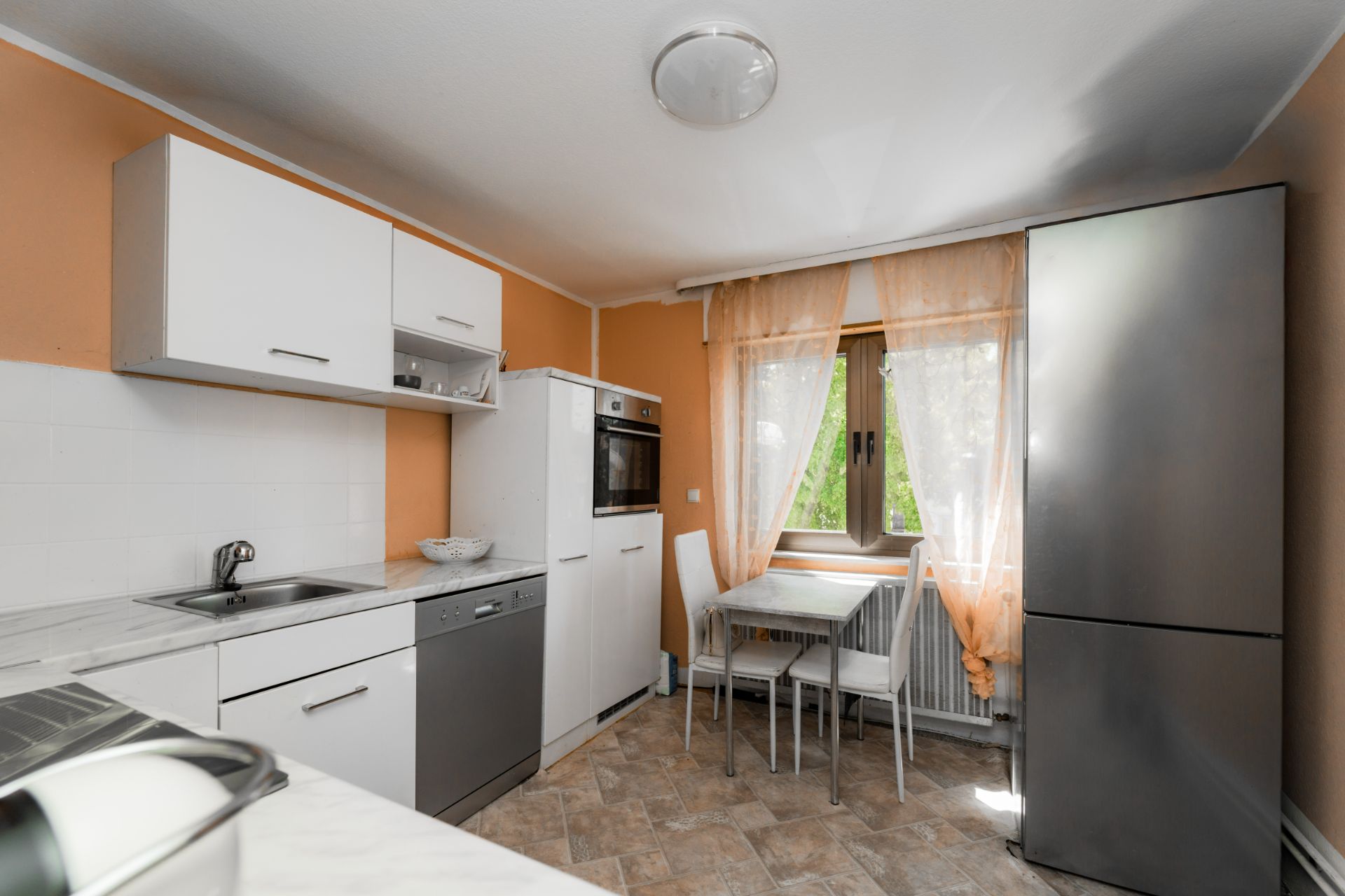 Küche mit weißer Einrichtung, Esstisch am Fenster und großem Kühlschrank.