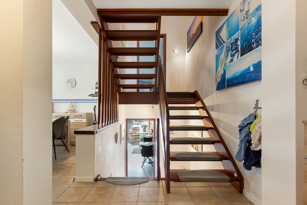 Holztreppe in einem Flur, Wände mit maritimen Bildern, Garderobe mit Kleidung rechts.