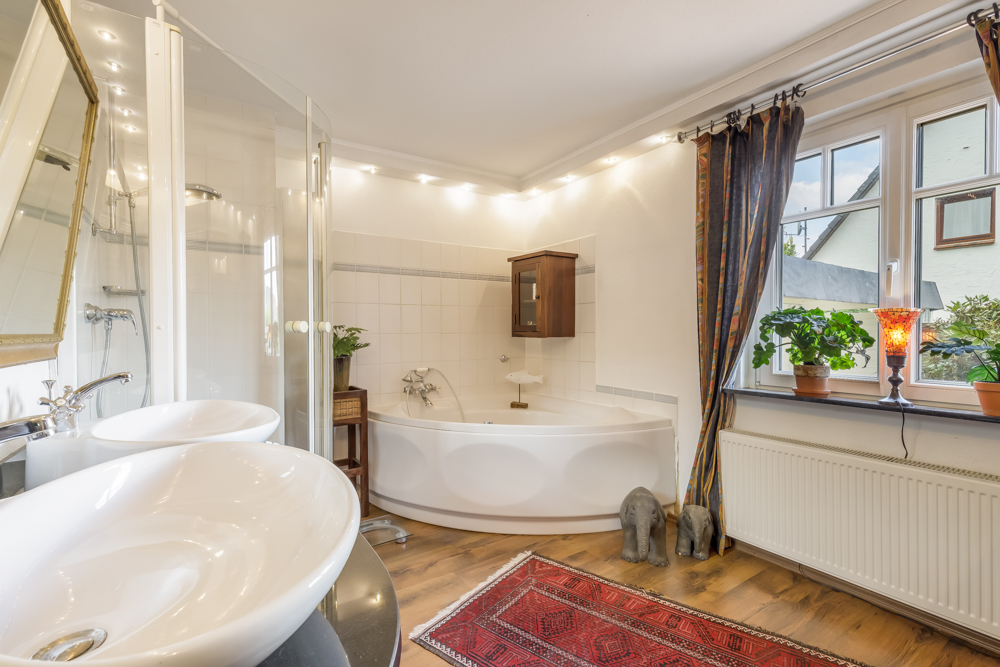 Helles Badezimmer mit Eckbadewanne, zwei Waschbecken, Dusche, Pflanzen und großem Fenster.