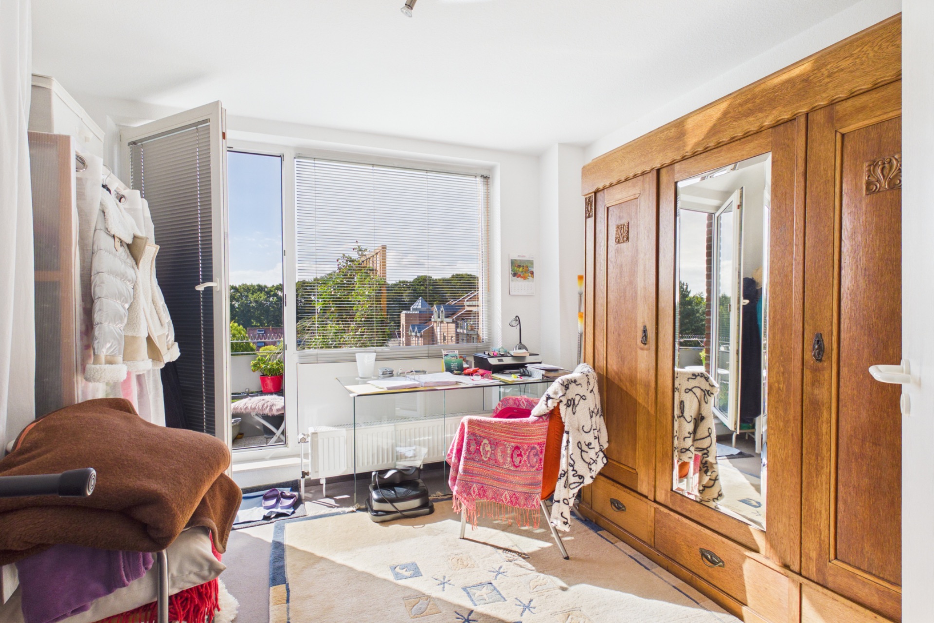 Helles Zimmer mit großem Holzschrank, Schreibtisch am Fenster und Zugang zu einem Balkon.