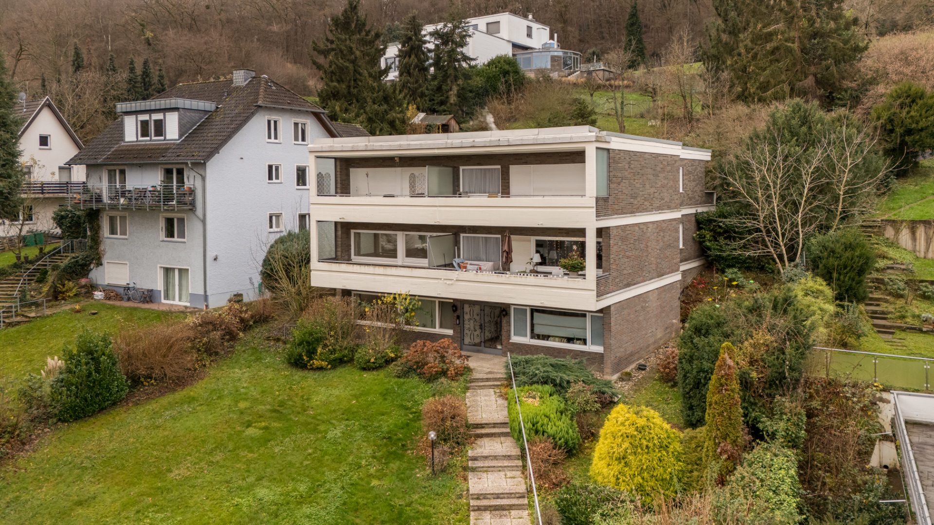Modernes dreistöckiges Wohnhaus mit Balkonen und Garten, eingefasst von Bäumen und Sträuchern.