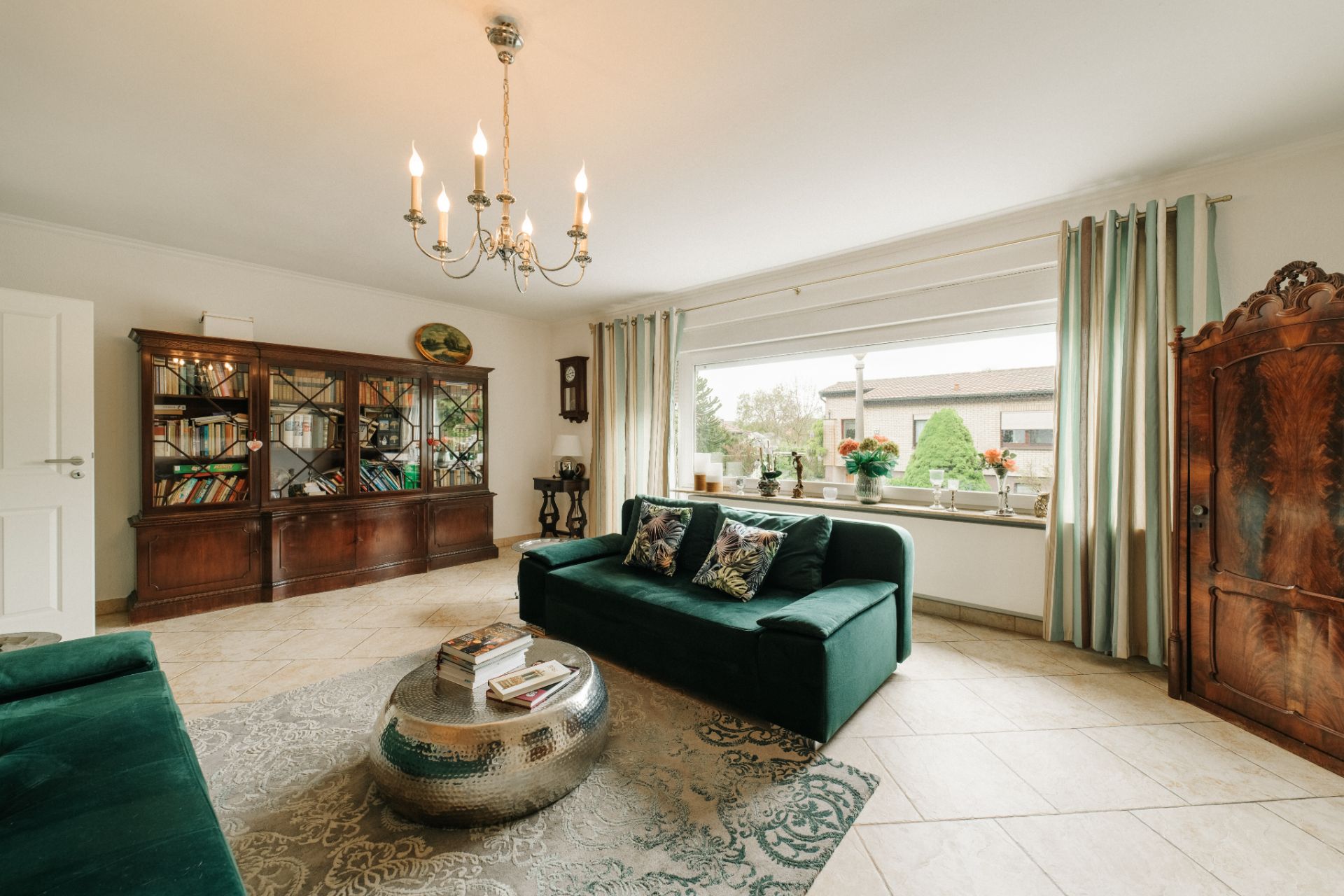 Helles Wohnzimmer mit grünem Sofa, Bücherregal, großem Fenster und Kronleuchter.