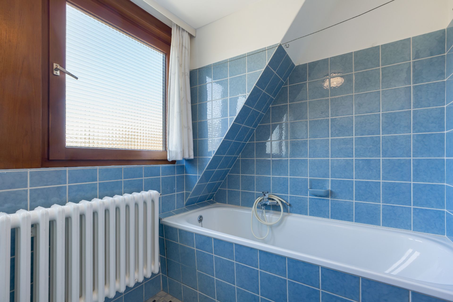 Badezimmer mit blauen Fliesen, Badewanne, Heizkörper und Fenster mit Vorhang.