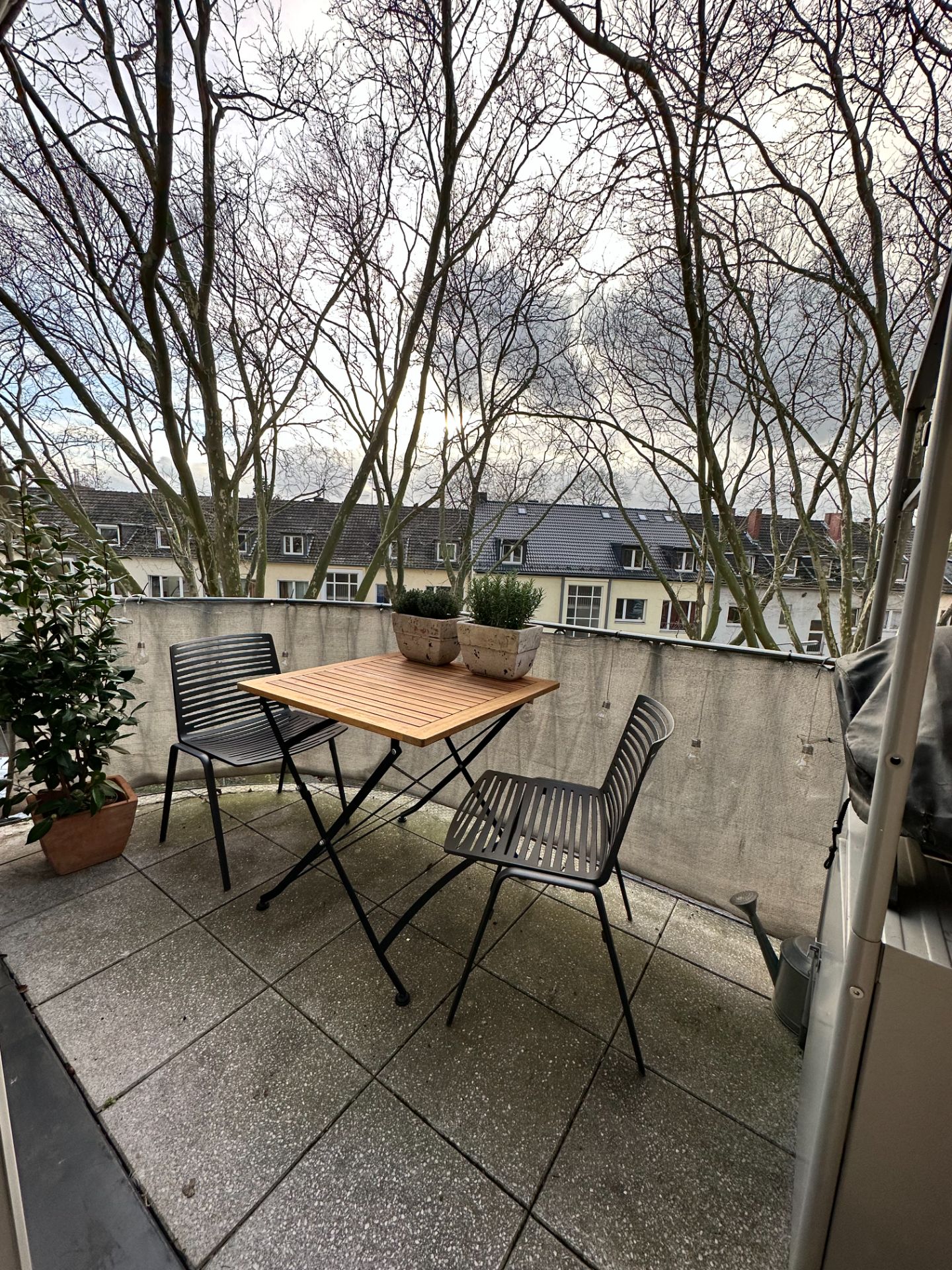 Balkon mit Tisch, zwei Stühlen, Pflanzkübeln und Blick auf Bäume und Häuser.