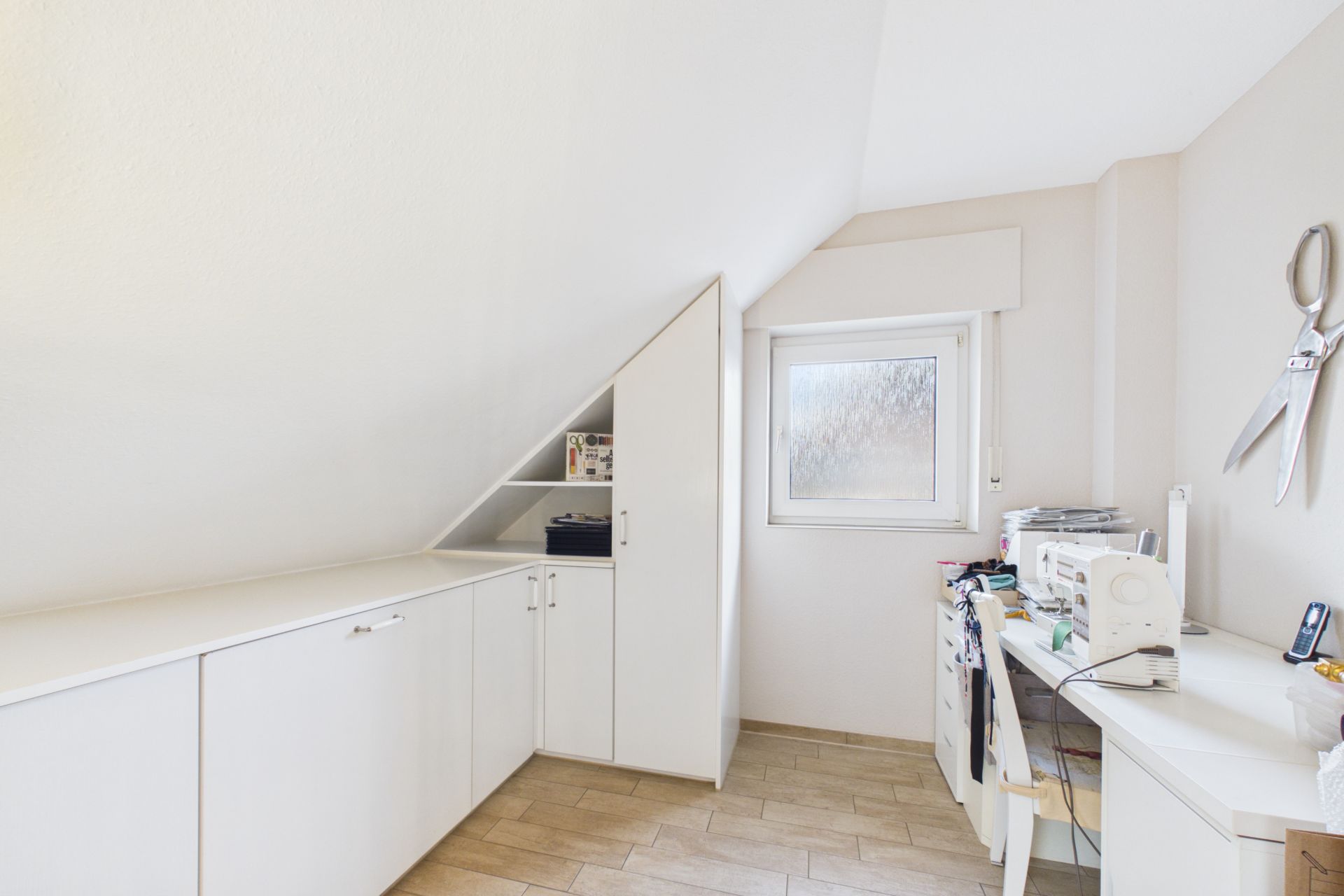 Helles Arbeitszimmer mit weißen Einbauschränken, einem Fenster und Schreibtisch unter einer Dachschräge.