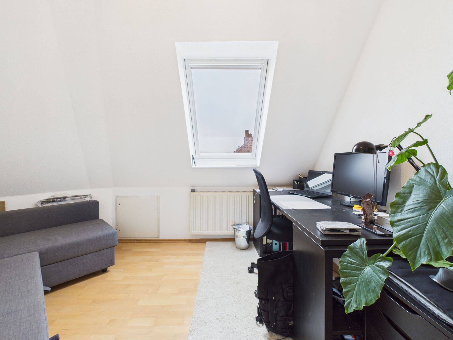 Helles Arbeitszimmer mit Dachfenster, Schreibtisch, Monitor, Pflanze und einem grauen Sofa.