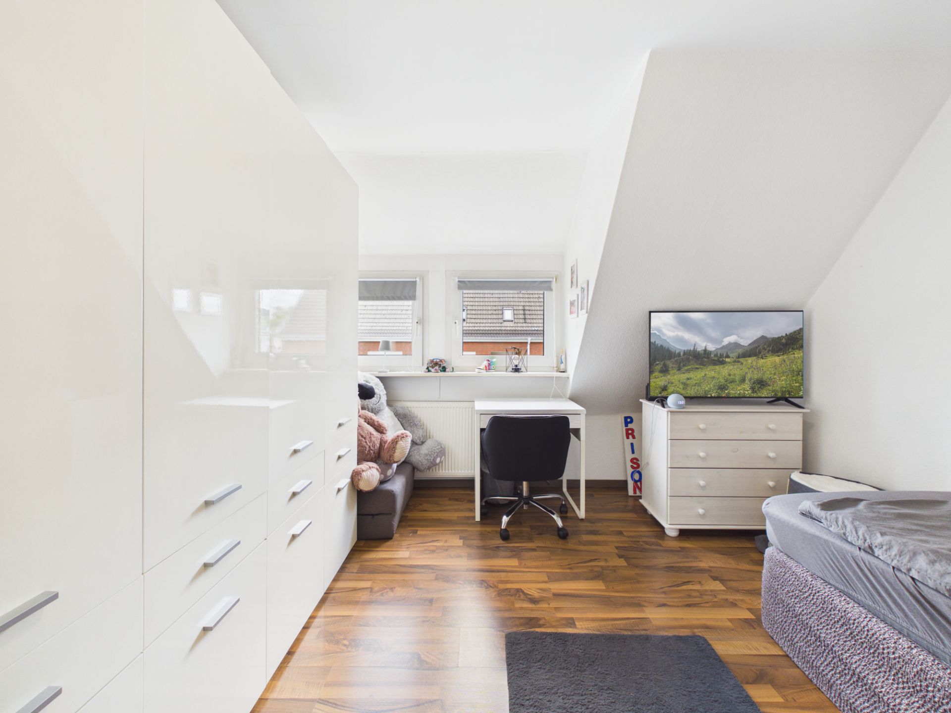 Helles Zimmer mit Kleiderschrank, Schreibtisch, Bett und Kommode mit Fernseher.