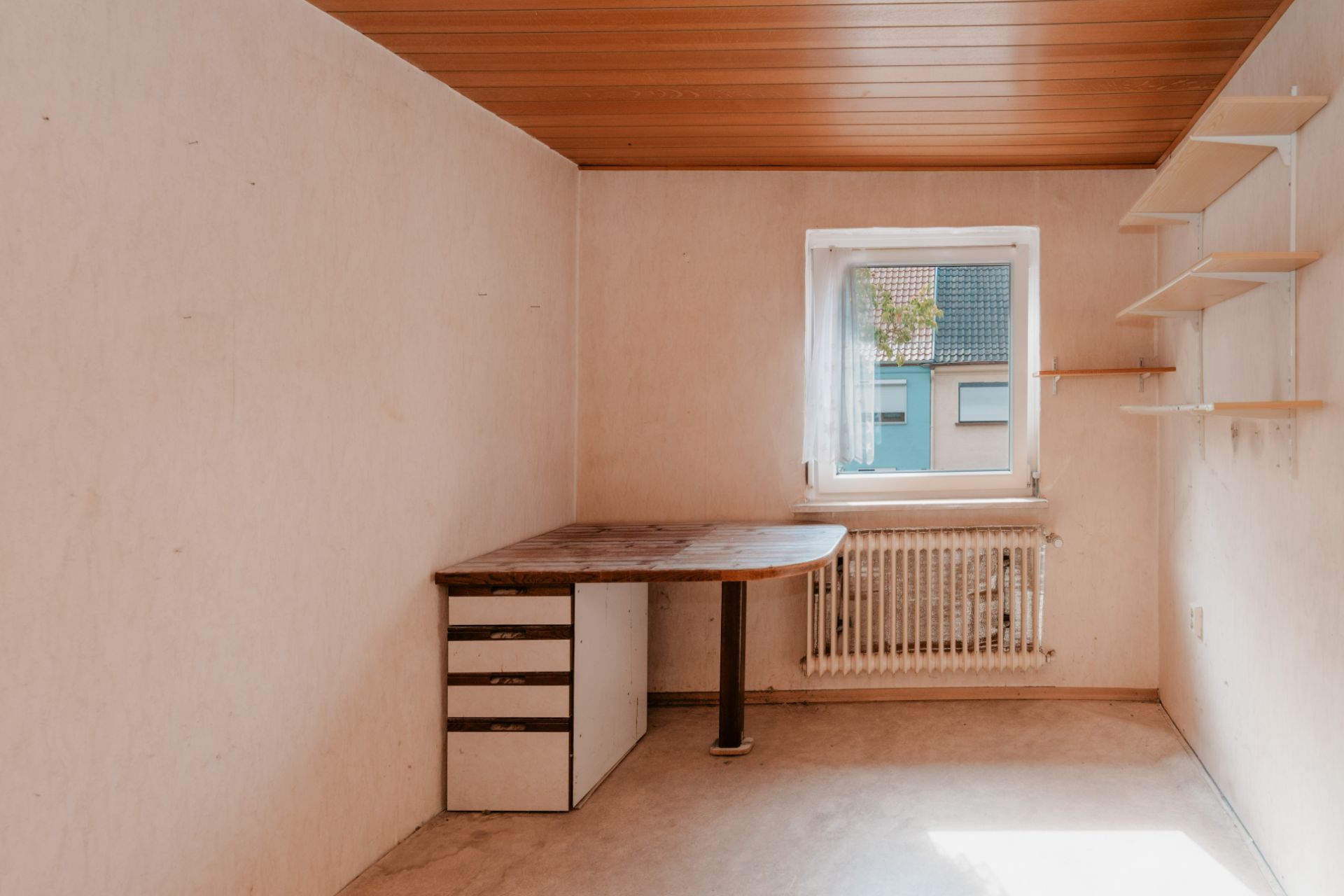 Kleines Zimmer mit Holztisch, Regalen an der Wand und Fenster mit Blick nach draußen.