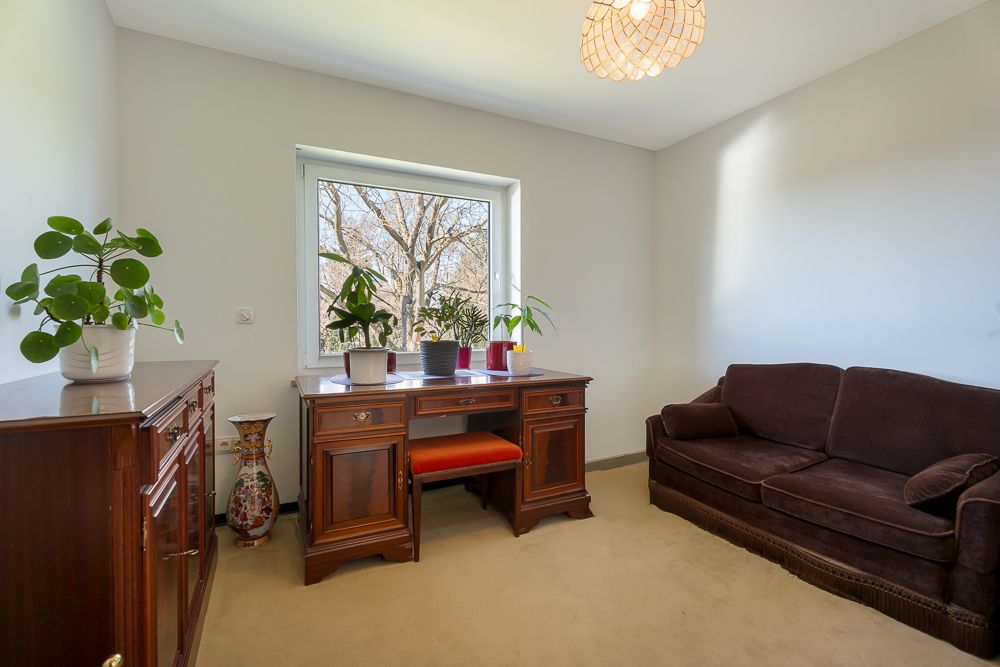 Helles Zimmer mit braunem Sofa, Holzschreibtisch, Pflanzen und Kommode am Fenster.