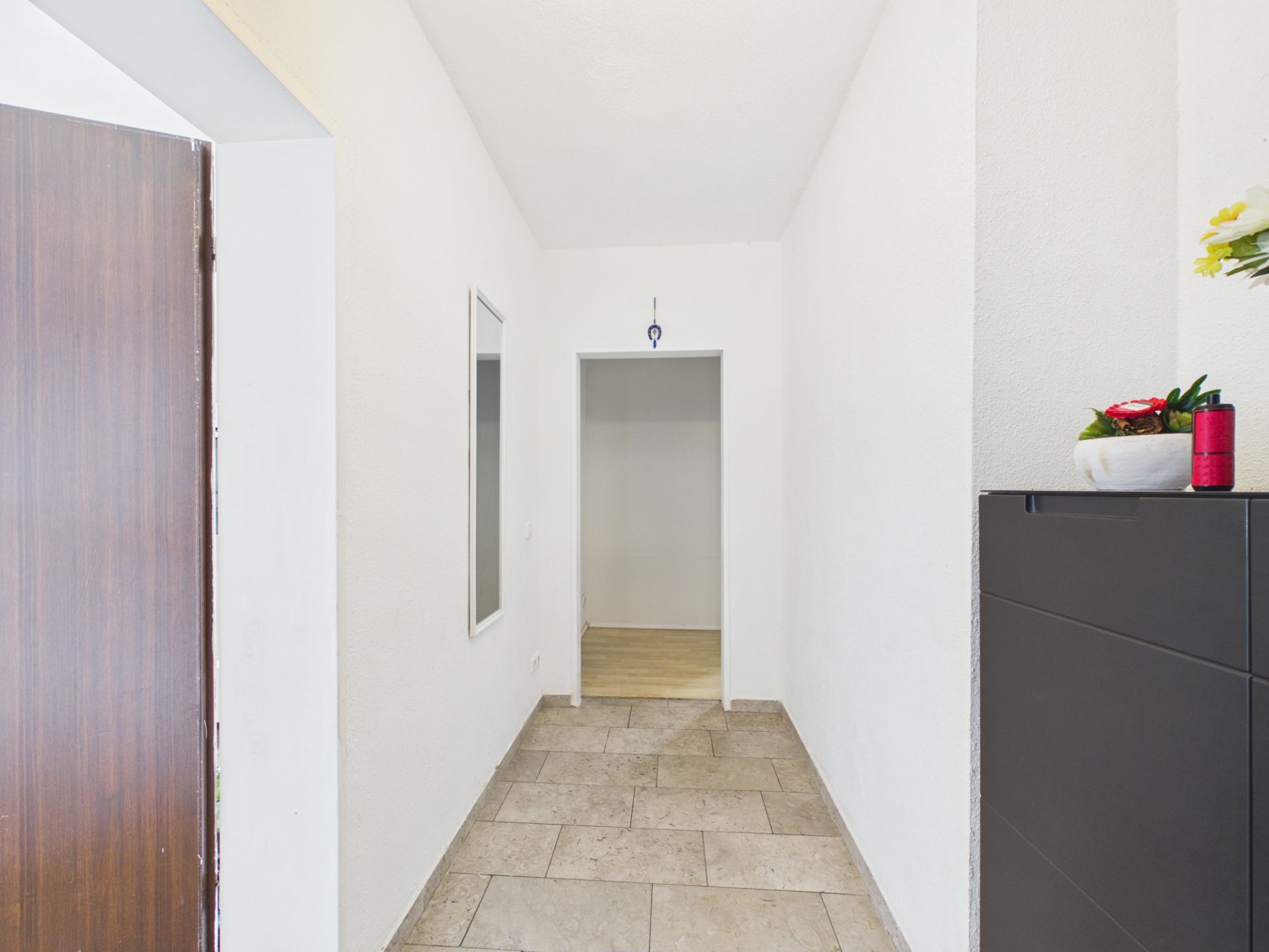 Heller Flur mit Fliesenboden, Spiegel an der Wand und Kommode mit Blumen.