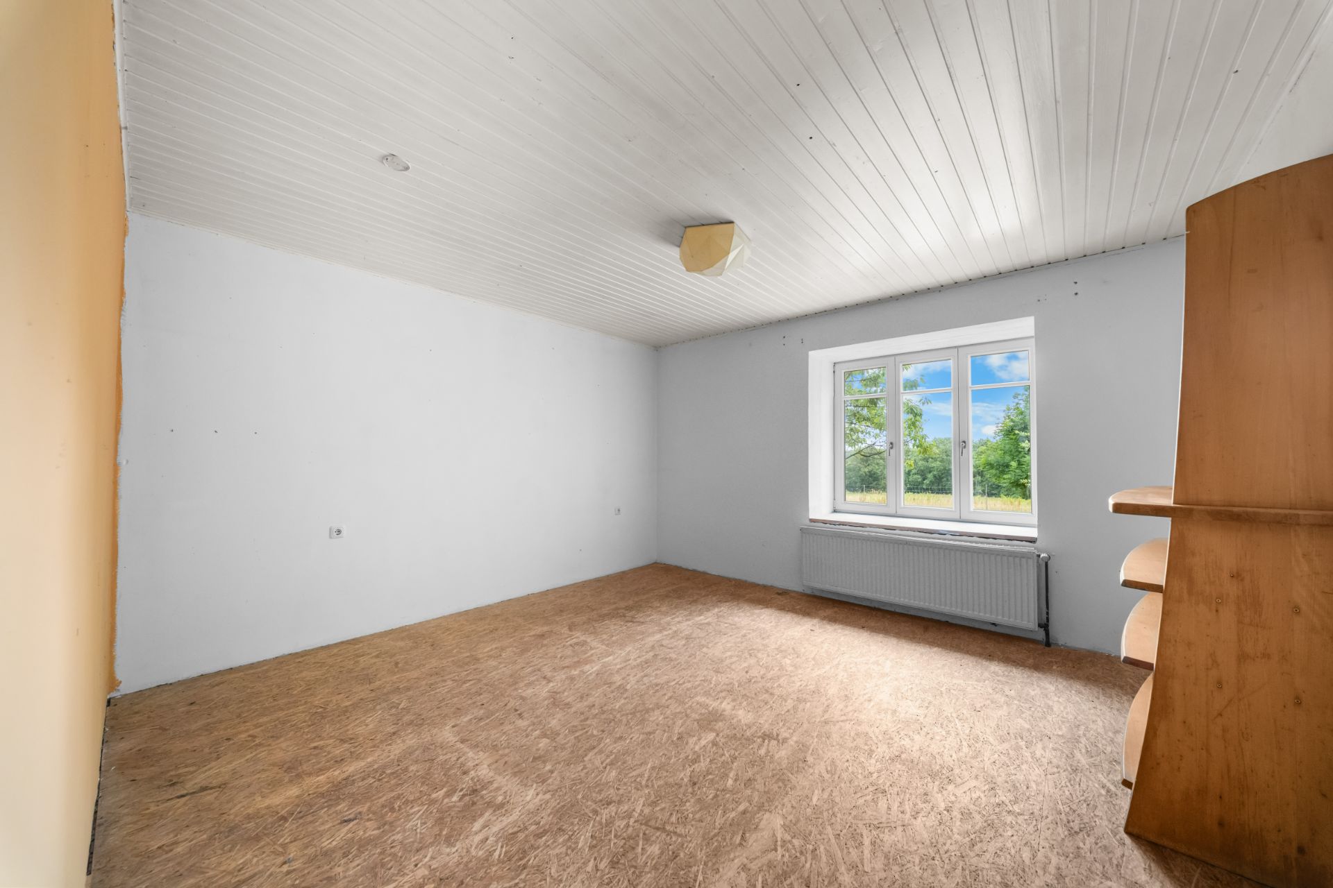 Leeres Zimmer mit großem Fenster, Heizkörper und Holzregal an der rechten Wand.