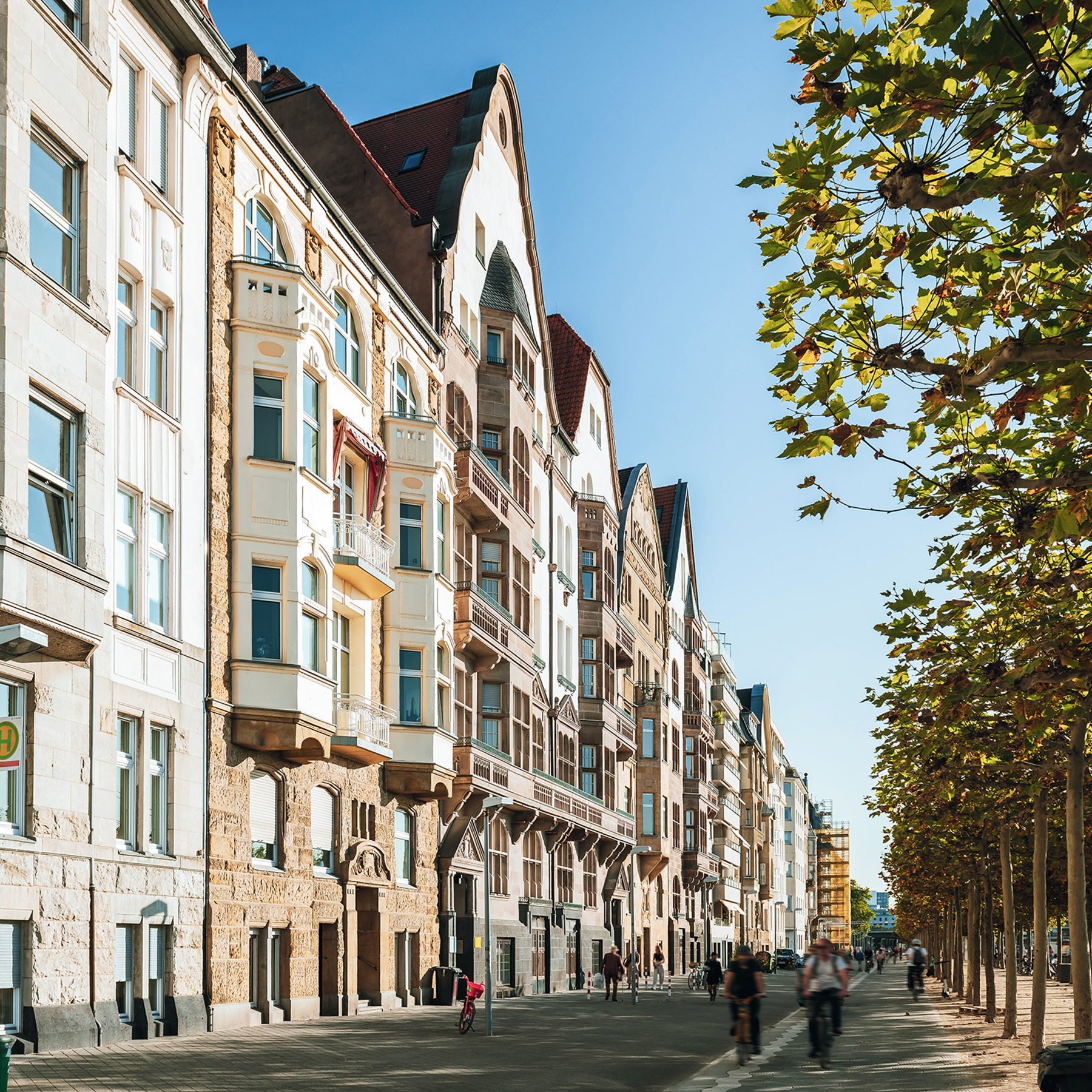Mehrstöckige Wohnhäuser an einer Straße mit Bäumen und Menschen auf dem Gehweg.