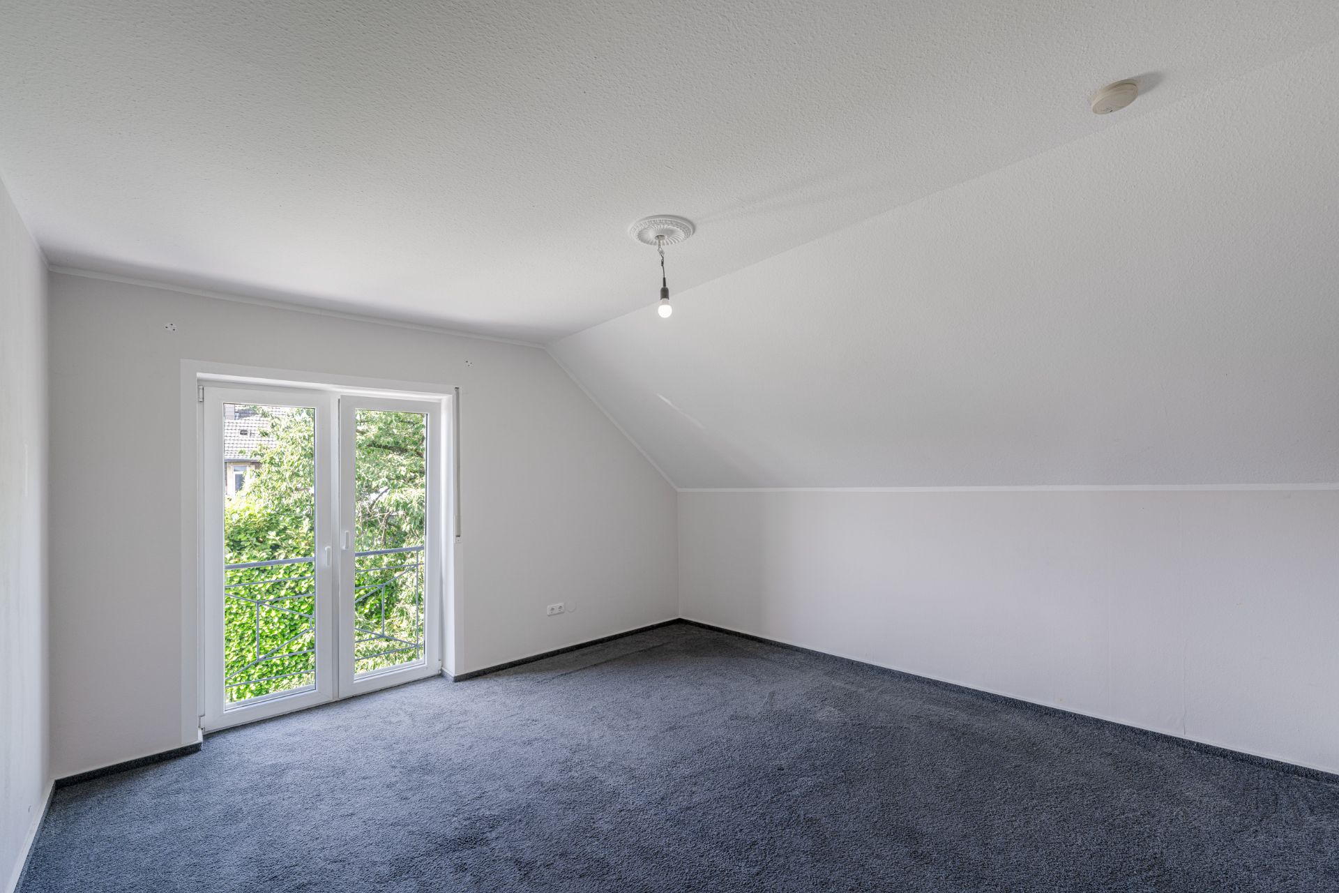 Leeres Zimmer mit Teppichboden, weißer Wand und Doppeltür zu einem kleinen Balkon.