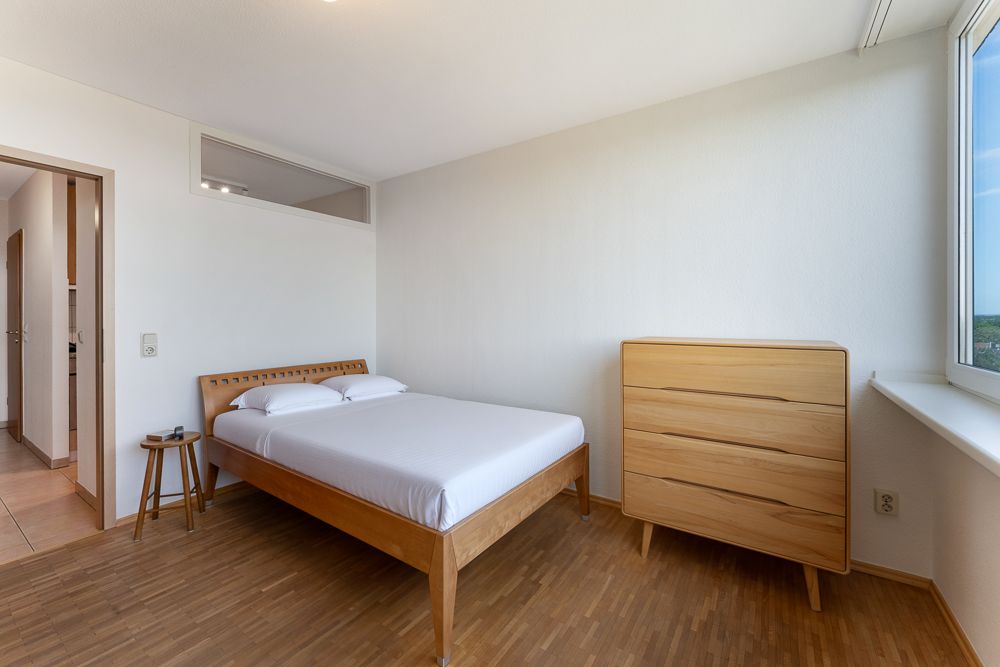 Helles Schlafzimmer mit Holzbett, Kommode und großem Fenster.