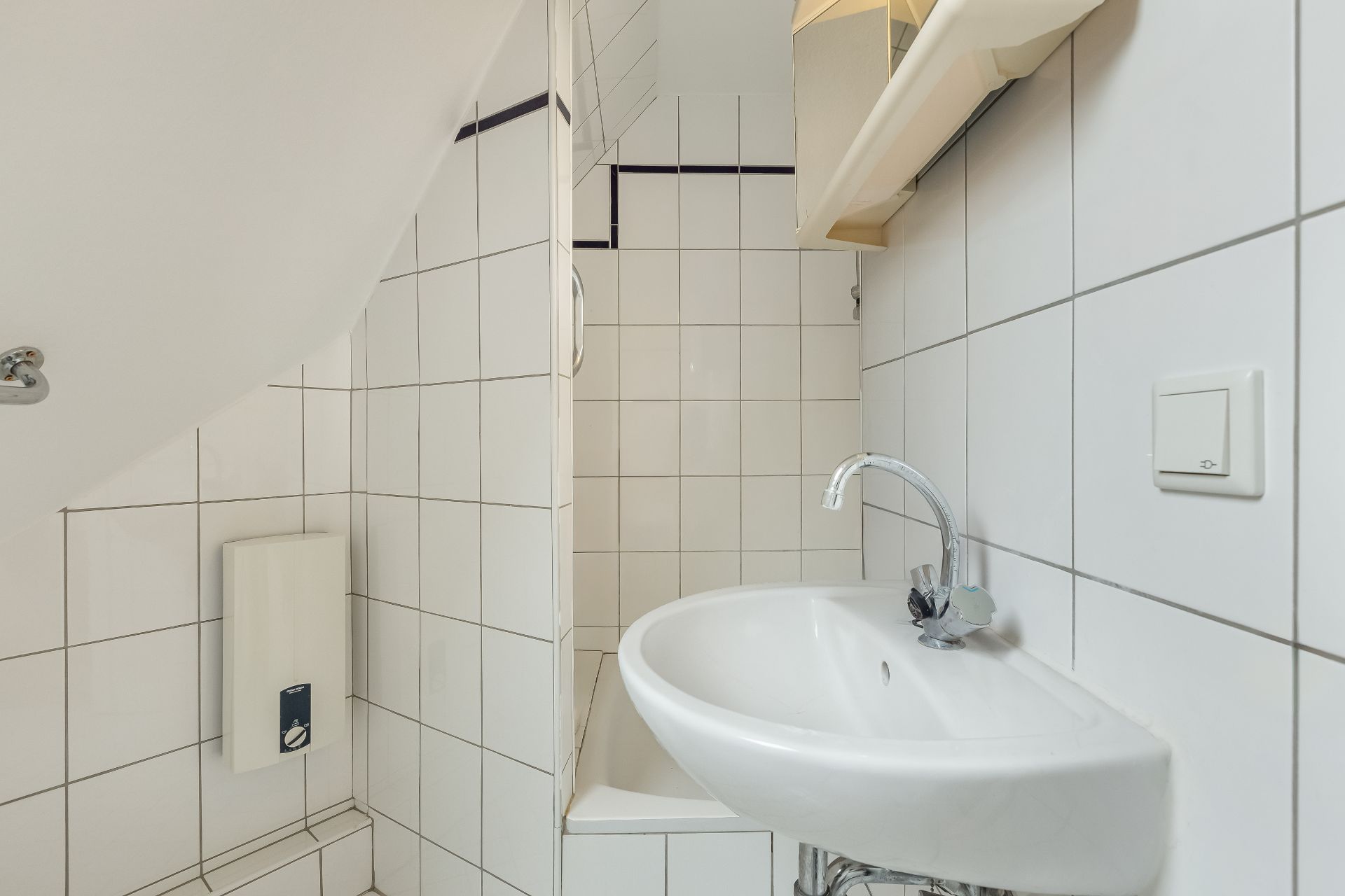 Kleines, weiß gefliestes Badezimmer mit Waschbecken und einem Wasserboiler an der Wand.