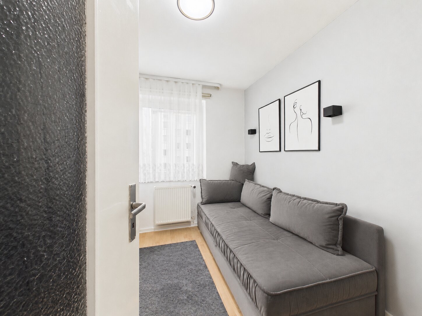 Kleines Zimmer mit grauem Sofa, Teppich, Fenster und zwei Bildern an der Wand.
