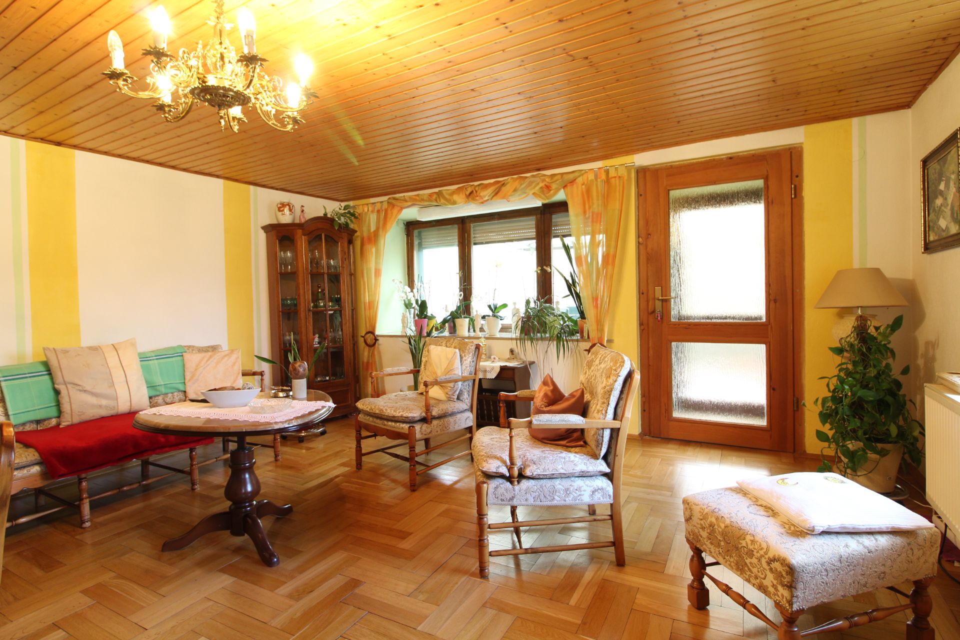 Helles Wohnzimmer mit Holzboden, Sesseln, Sofa, Holztisch und großen Fenstern mit Vorhängen.