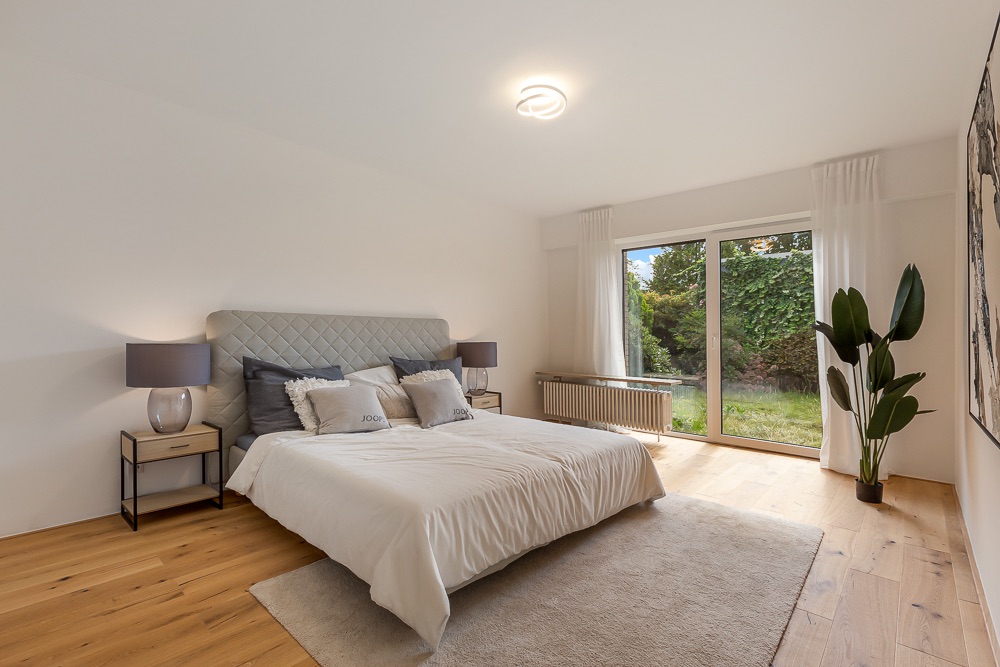 Modernes Schlafzimmer mit Doppelbett, Nachttischen, großen Fenstern und Blick auf den Garten.