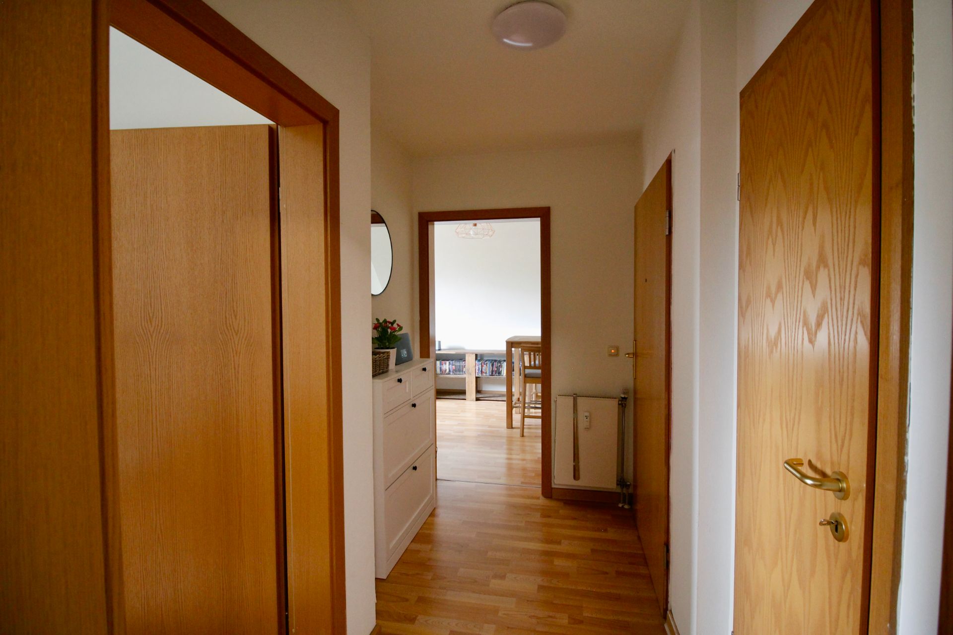 Flur mit Holzboden, weißen Wänden und Holztüren, Blick ins Wohnzimmer mit Möbeln.