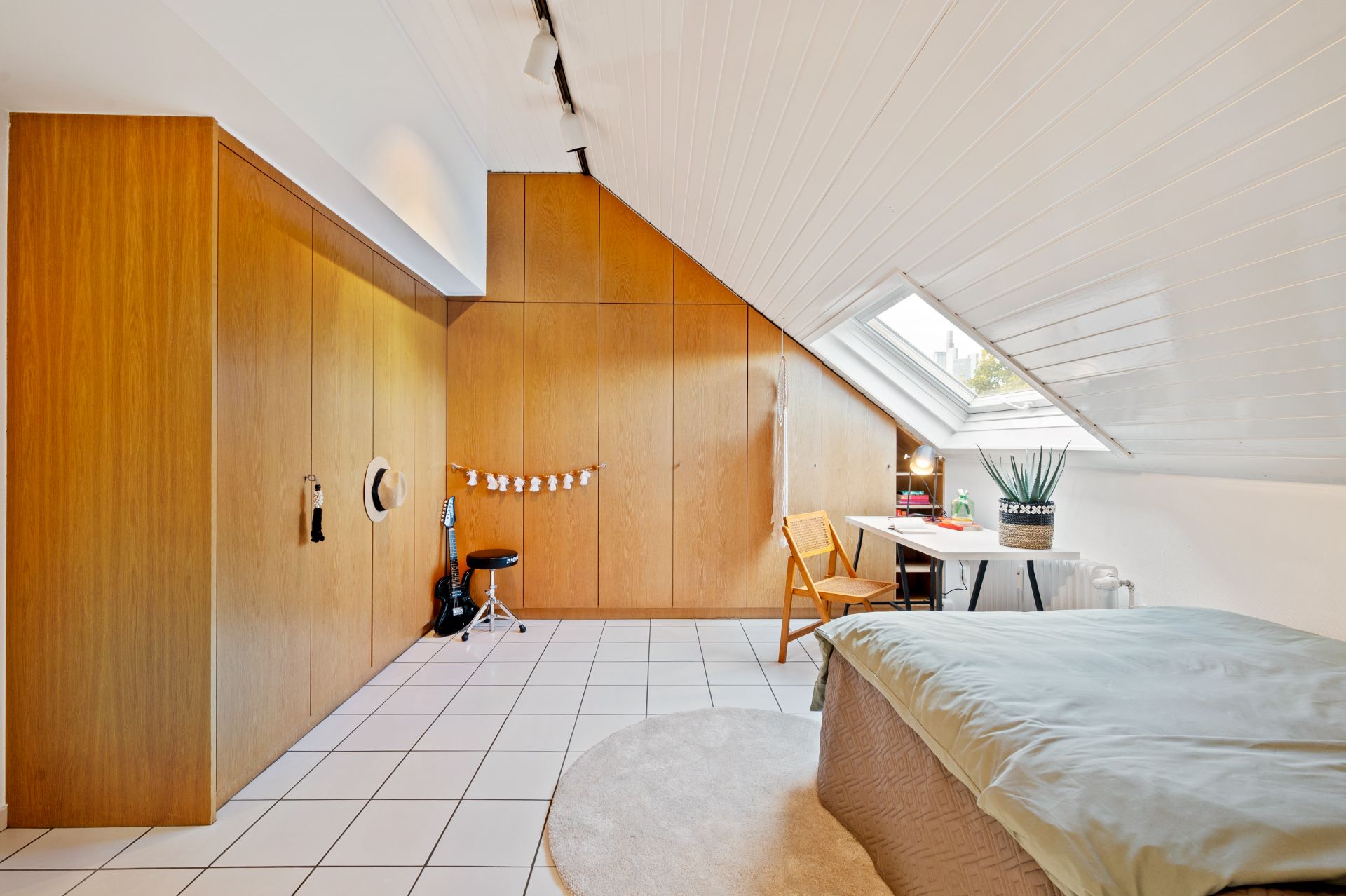 Helles Dachzimmer mit Holzwänden, Bett, Schreibtisch am Fenster und Fliesenboden.