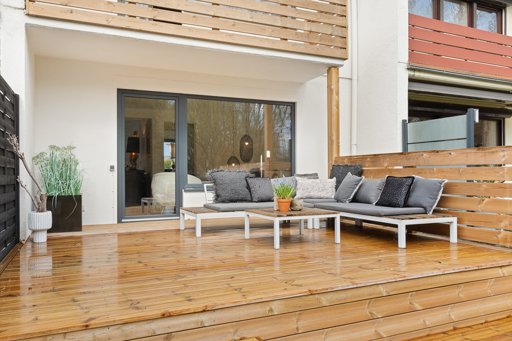 Holzterrasse mit grauem Sofa, Tisch, Pflanze und Holzzaun vor einer Glastür.