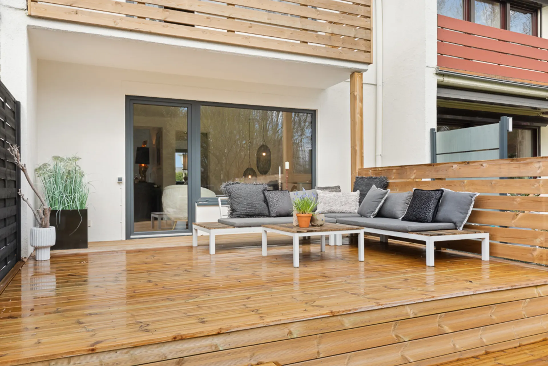Holzterrasse mit grauem Sofa, Tisch, Pflanze und Holzzaun vor einer Glastür.