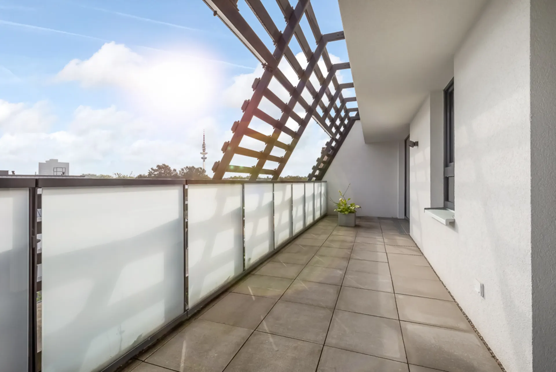 Großer Balkon mit Fliesenboden, Glasgeländer und Pflanze in einem Topf bei Sonnenschein.