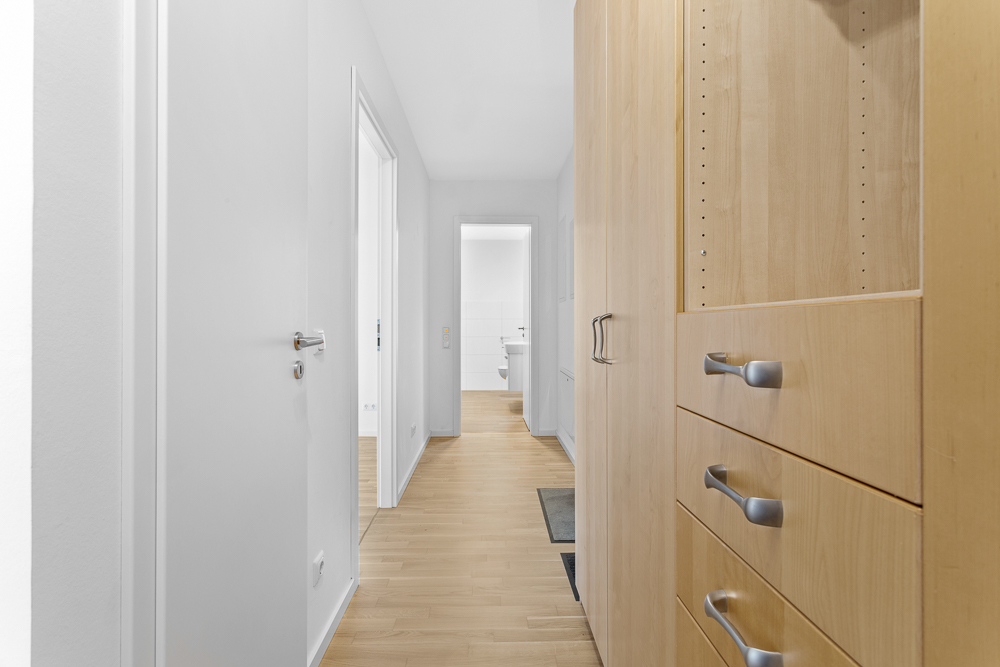 Heller Flur mit Holzboden, Holzschrank und Blick in ein weiß gefliestes Badezimmer.