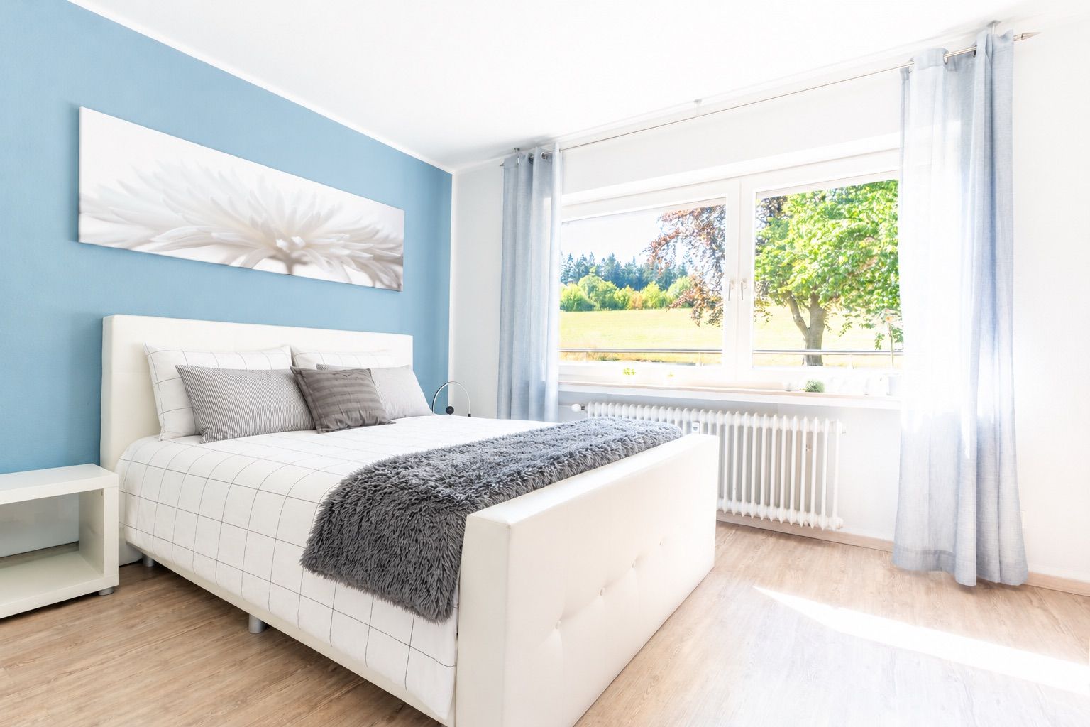 Helles Schlafzimmer mit weißem Bett, blauer Wand, großem Fenster und Blick ins Grüne.