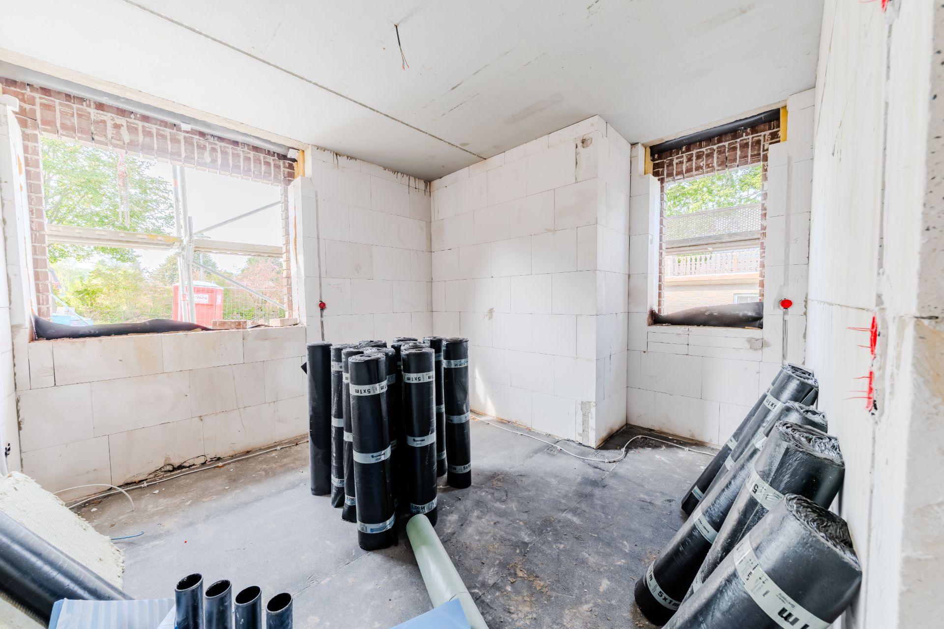 Heller Raum im Rohbau mit zwei Fenstern und Baumaterial auf dem Boden.