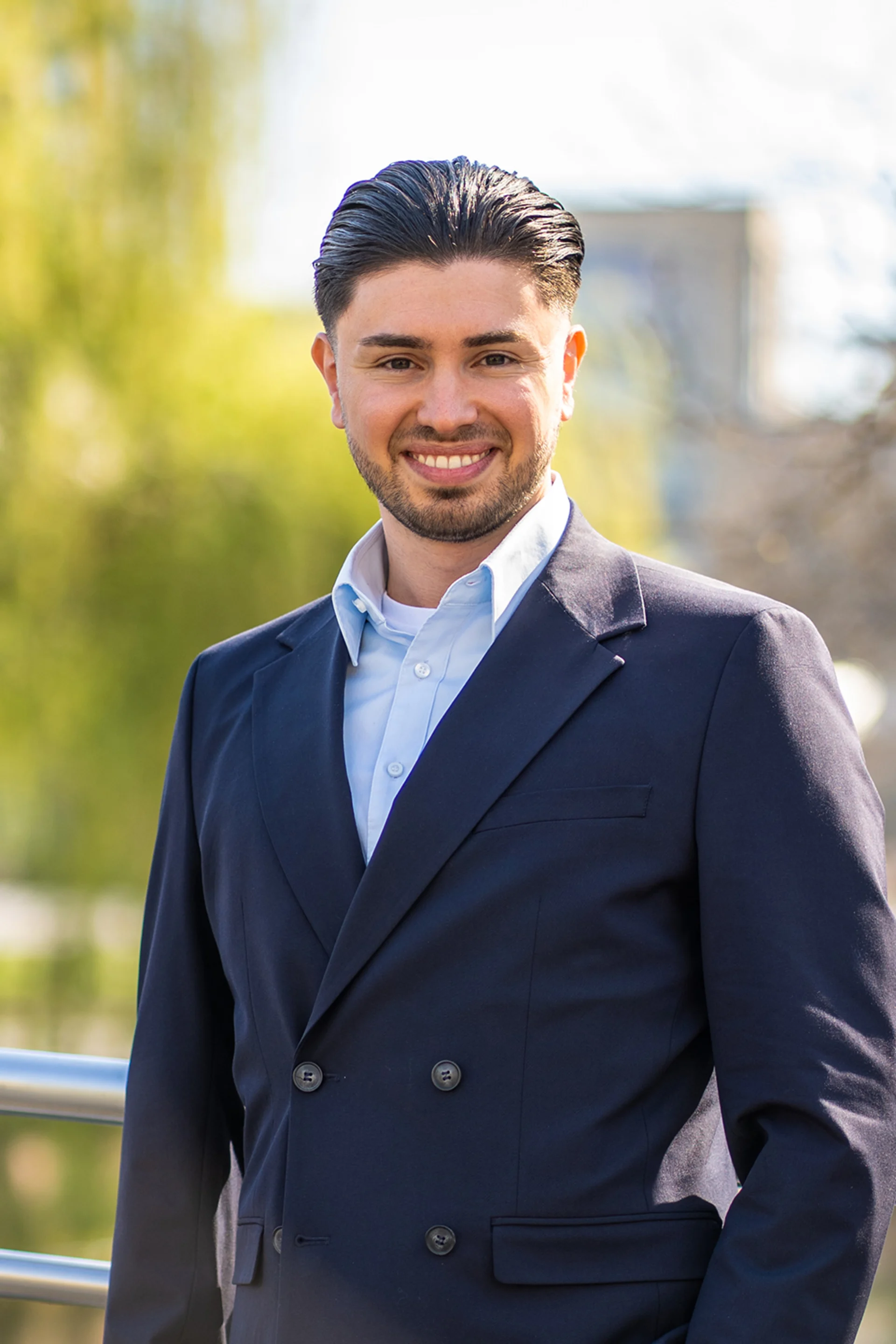 Ein Mann mit dunklem Haar im Anzug steht draußen vor grünen Bäumen.