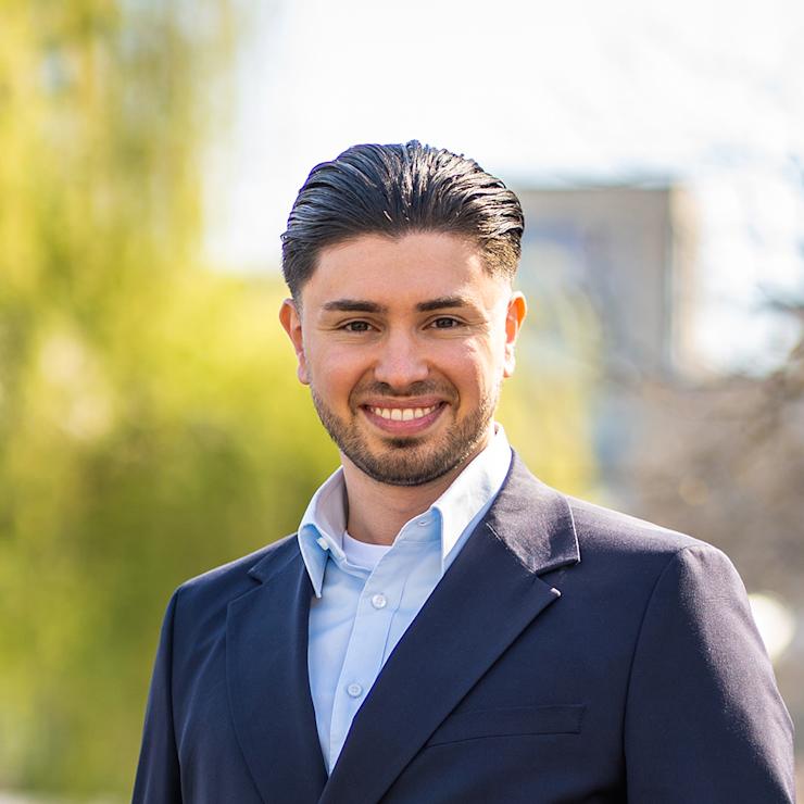 Ein Mann mit dunklem Haar im Anzug steht draußen vor grünen Bäumen.