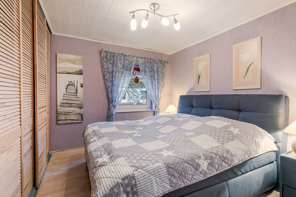 Schlafzimmer mit großem Bett, lilafarbener Tapete, Bildern an der Wand und Fenster mit Vorhang.