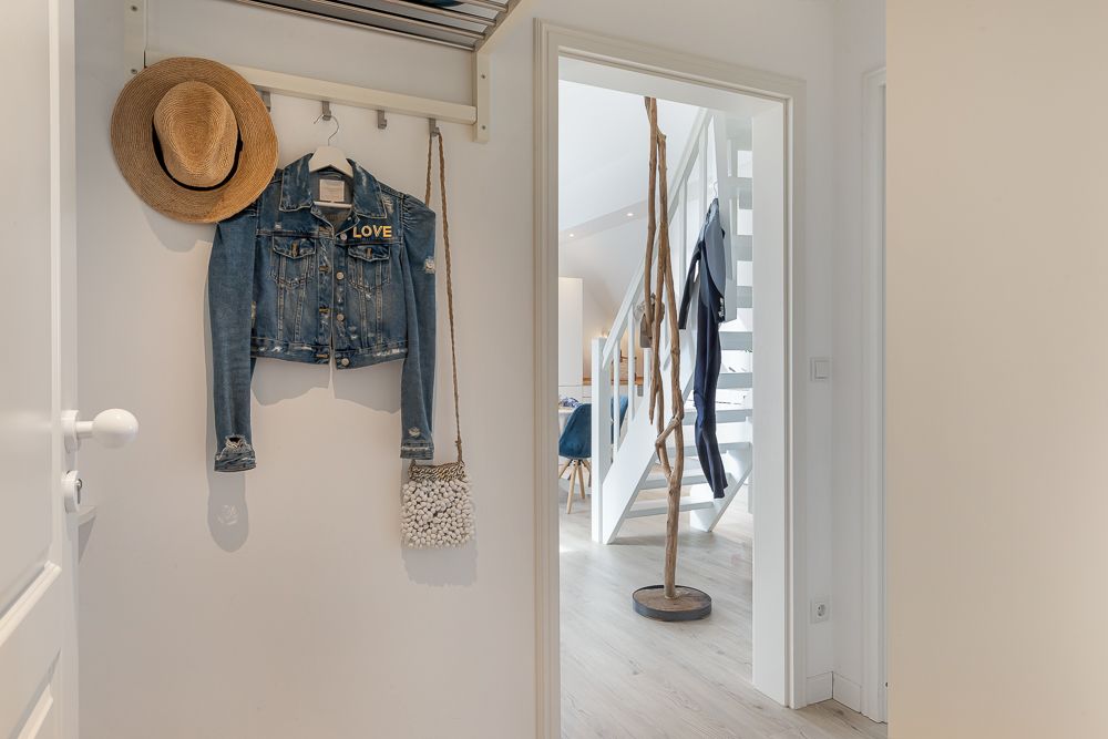 Eingangsbereich mit Hut, Jeansjacke und kleiner Tasche an einer Garderobe, Blick ins Wohnzimmer mit Treppe.