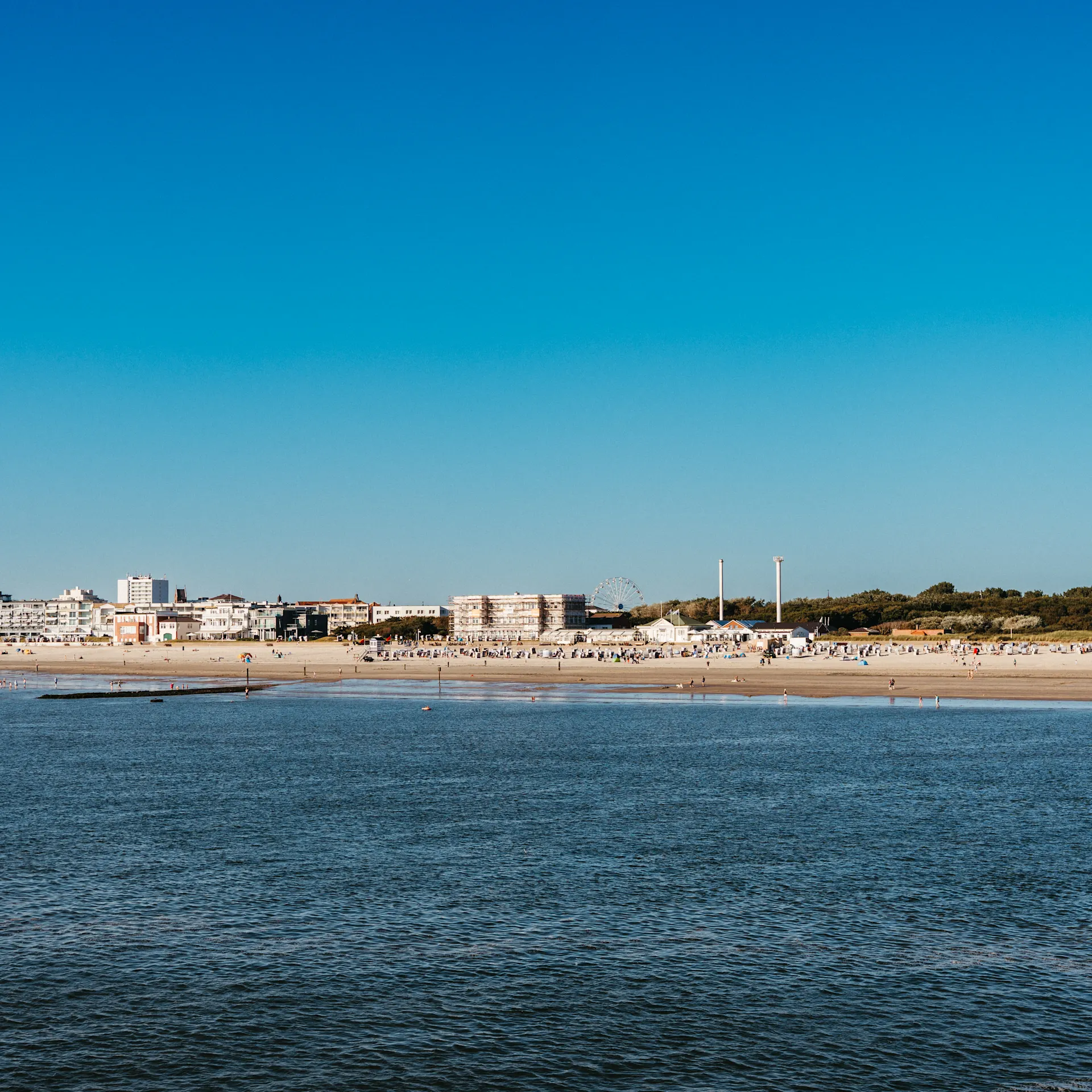 Blick auf einen Sandstrand mit Gebäuden im Hintergrund und blauem Himmel.