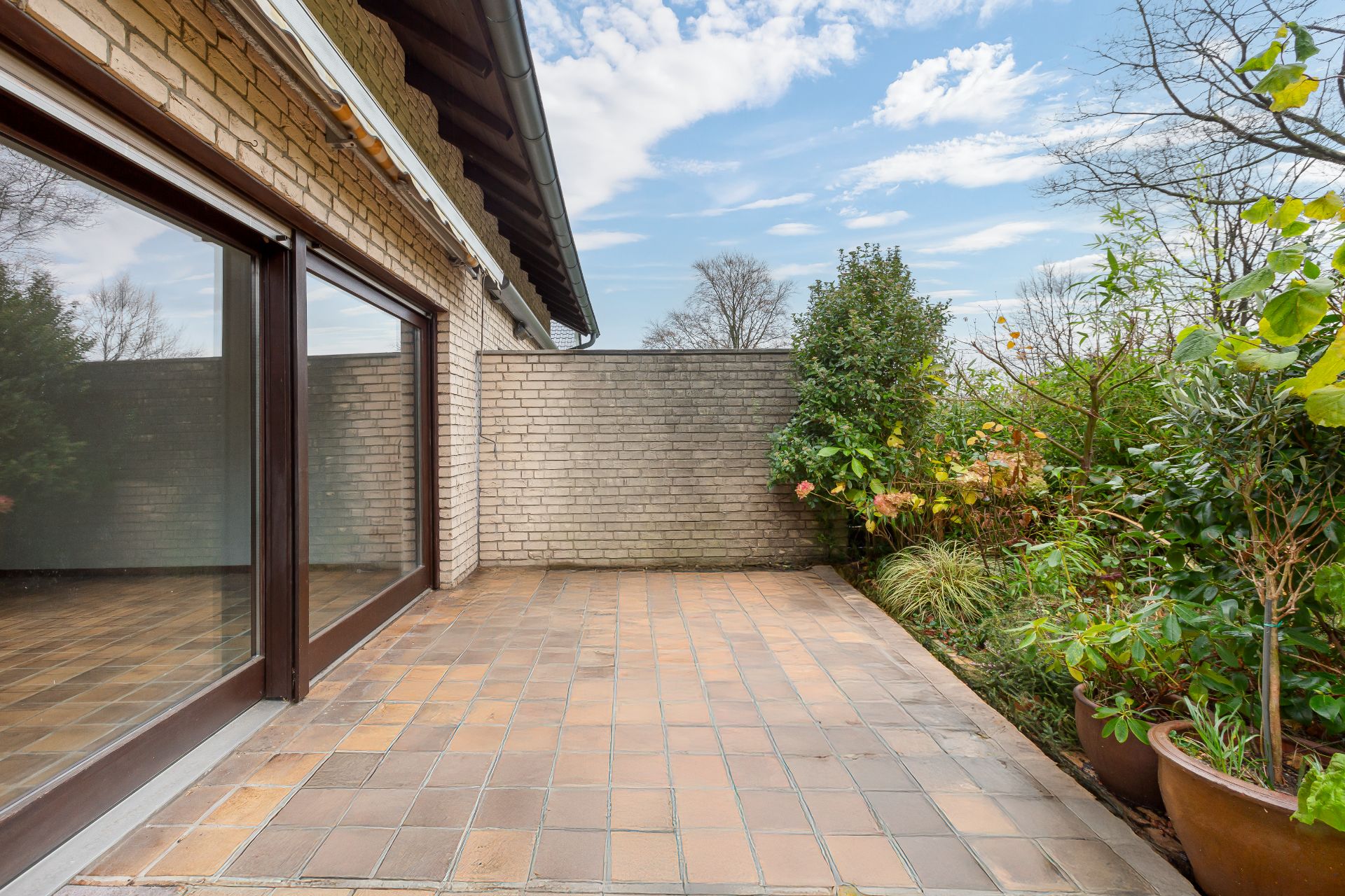 Geflieste Terrasse mit großen Fenstern, angrenzendem Mauerwerk und Pflanzen in Töpfen.