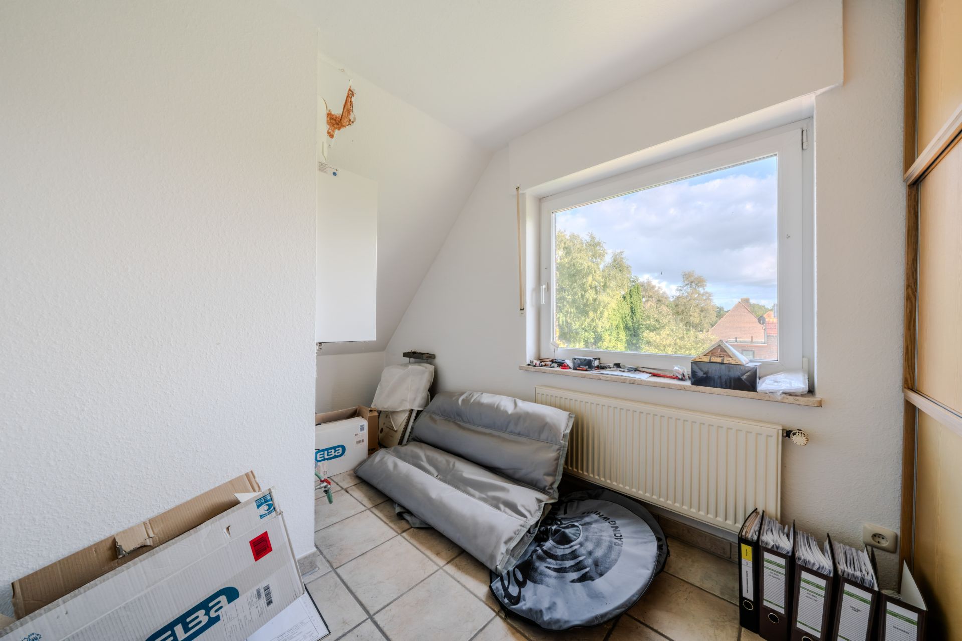 Kleiner Raum mit einem Fenster, Kartons, Ordnern und zusammengeklapptem Sitzpolster.