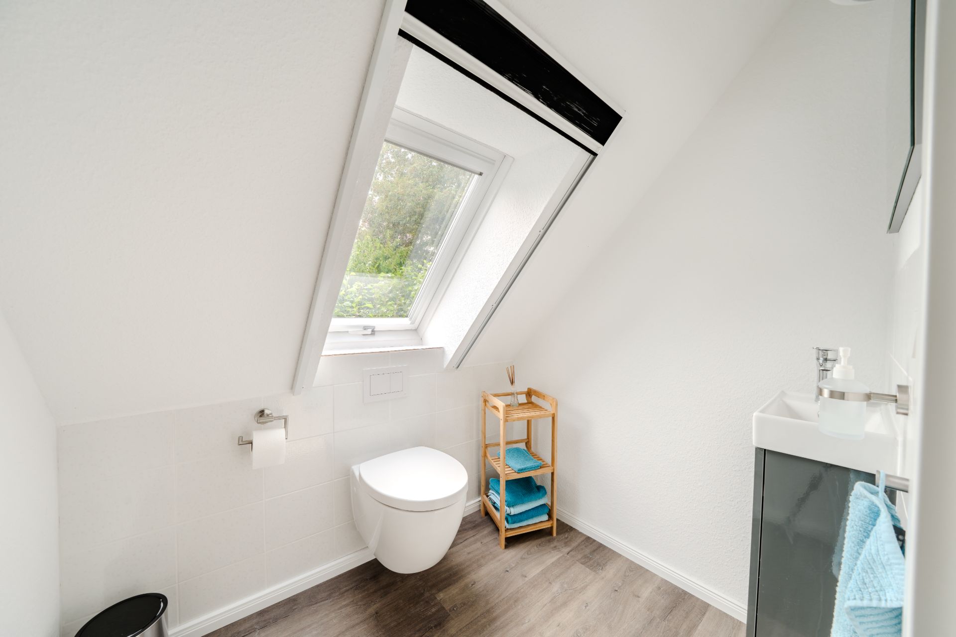 Helles Bad mit Dachschräge, Fenster, Wandtoilette, kleinem Waschbecken und Regal mit Handtüchern.