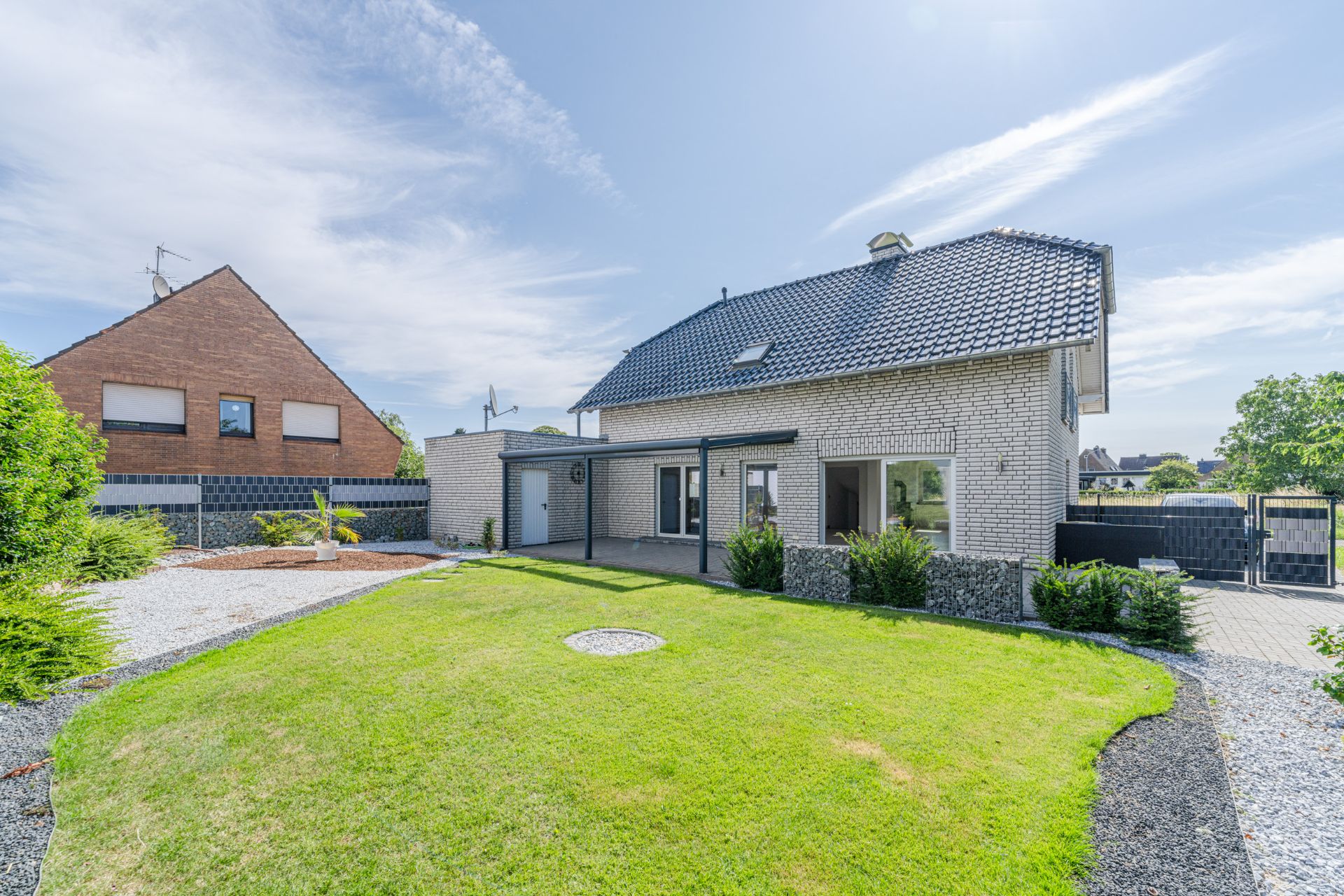 Weißes Einfamilienhaus mit Garten, Terrasse und bepflanzten Randsteinen bei sonnigem Wetter.