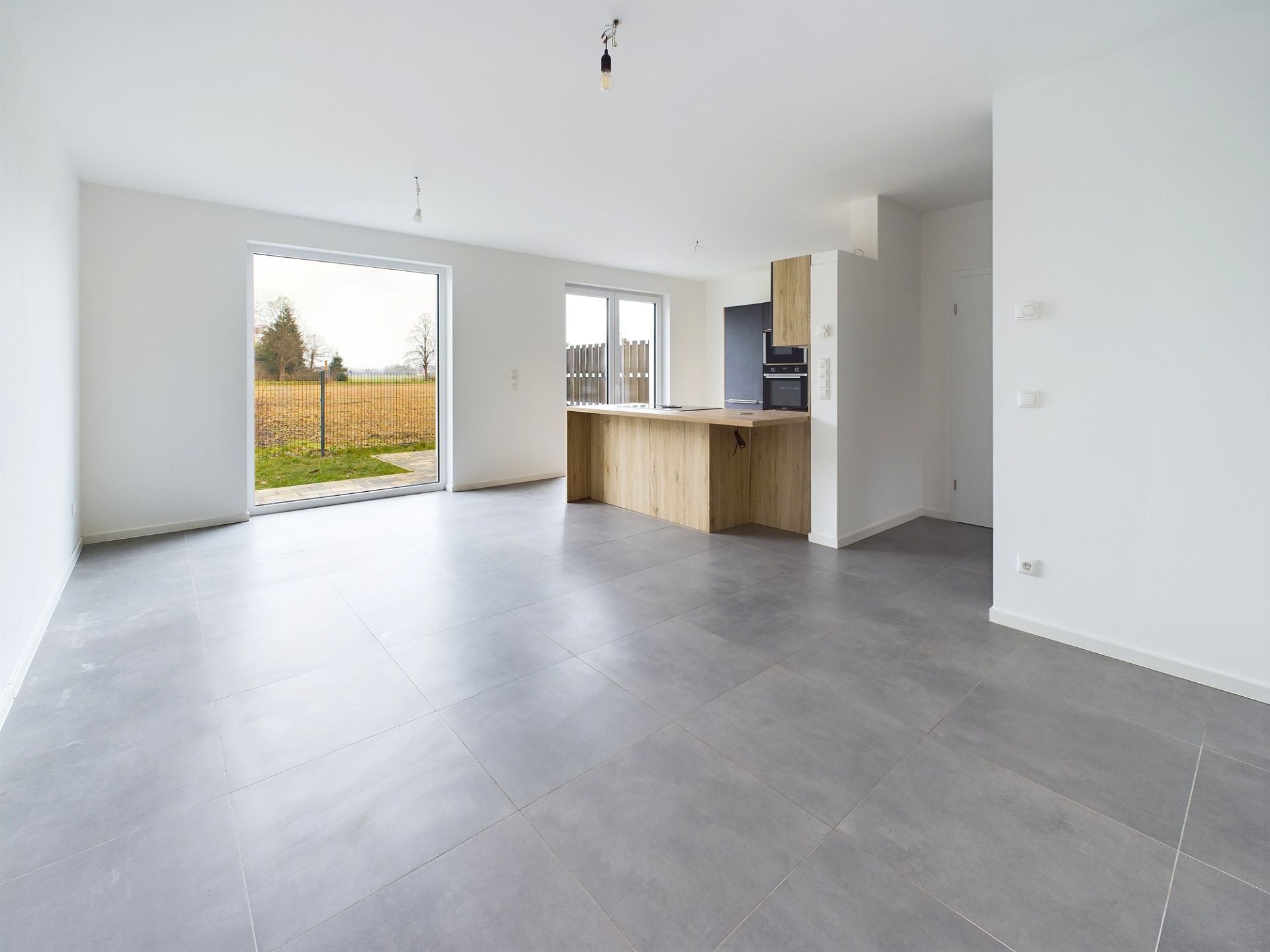 Leerstehender, heller Wohnraum mit großen Fenstern und moderner Einbauküche.