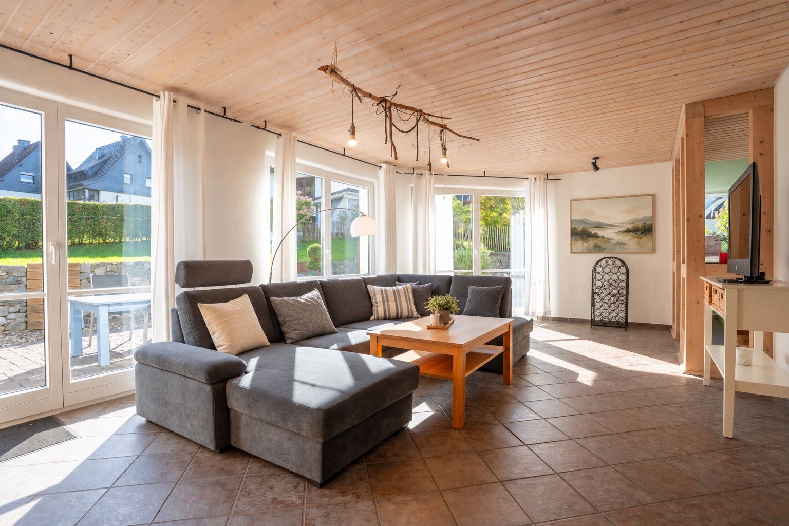 Helles Wohnzimmer mit großem grauen Sofa, Holztisch und großen Fenstern zur Terrasse.