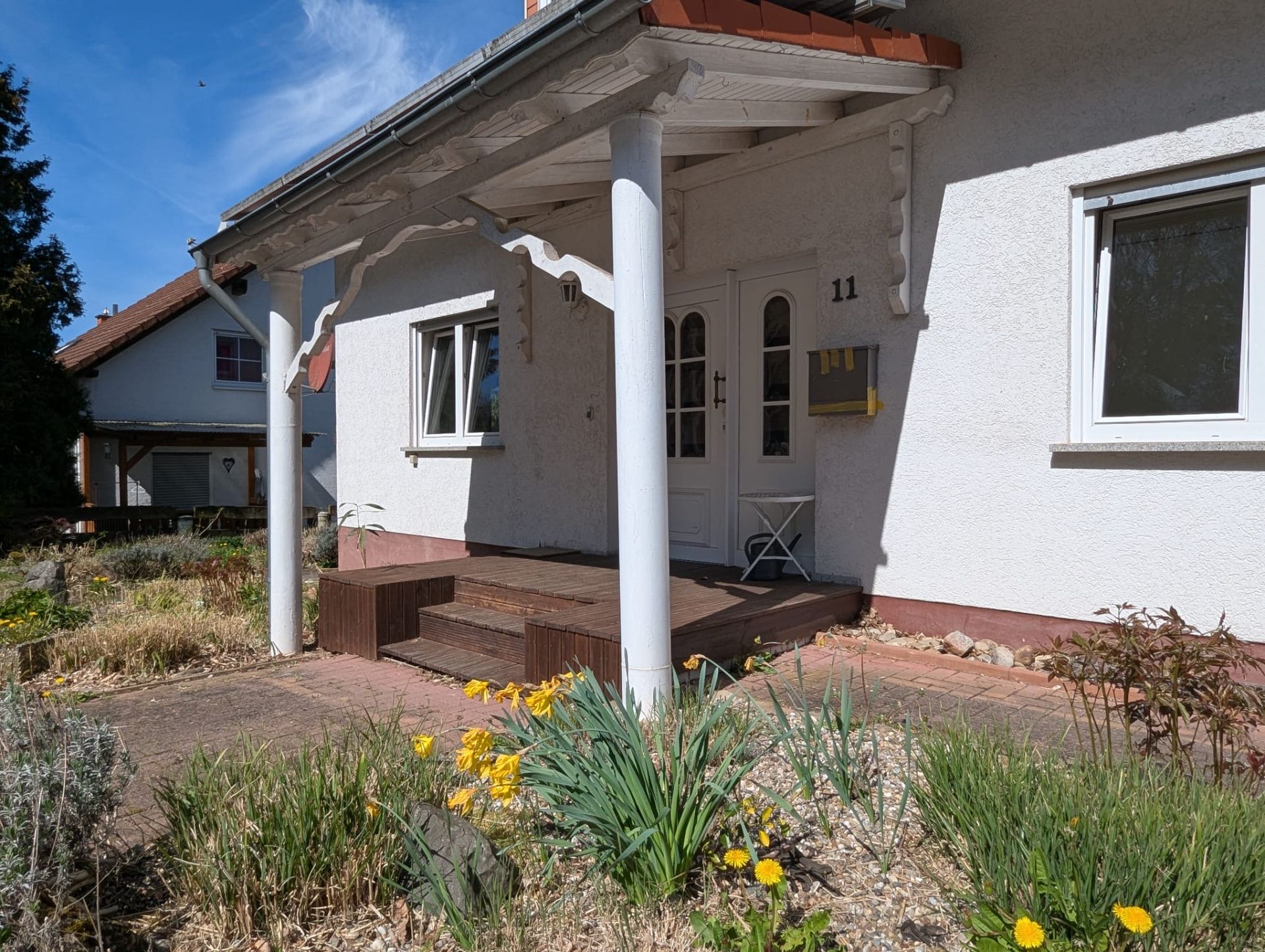 Eingangsbereich eines Hauses mit weißer Tür, Veranda und Garten mit gelben Blumen.
