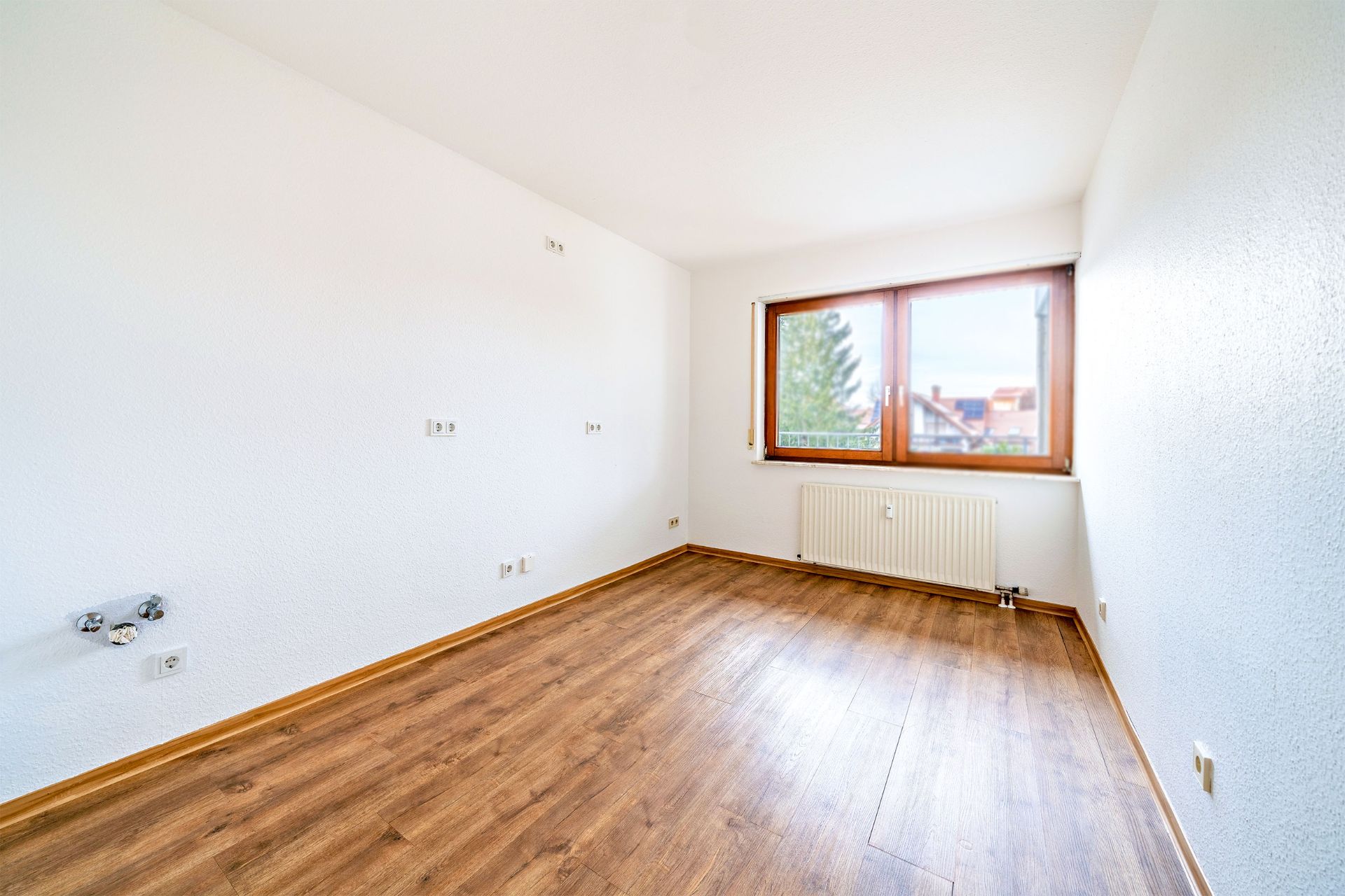 Heller, leerer Raum mit Holzfußboden, weißer Wand und großem Fenster.