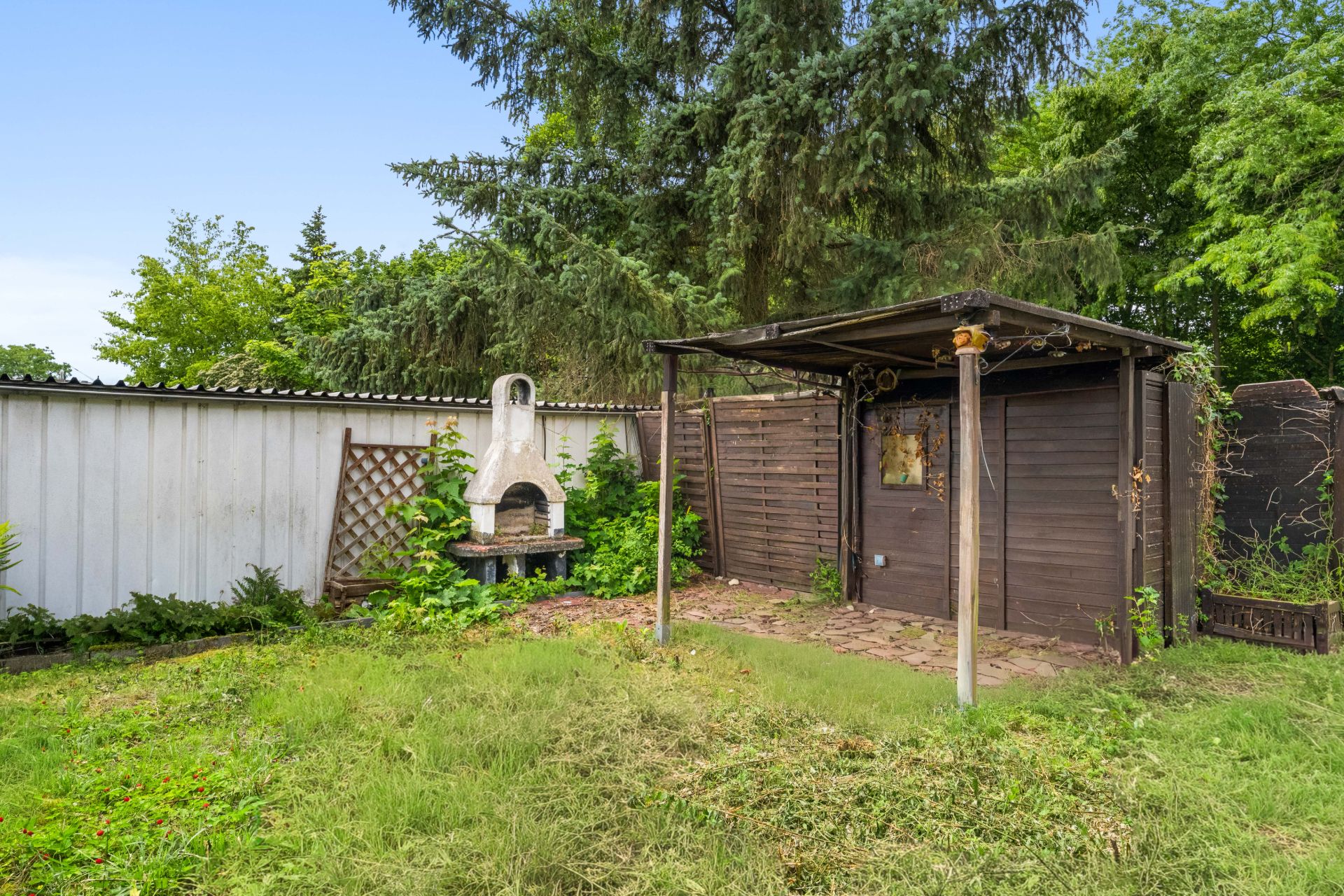 Garten mit Rasenfläche, Grill und überdachtem Holzschuppen vor Bäumen.