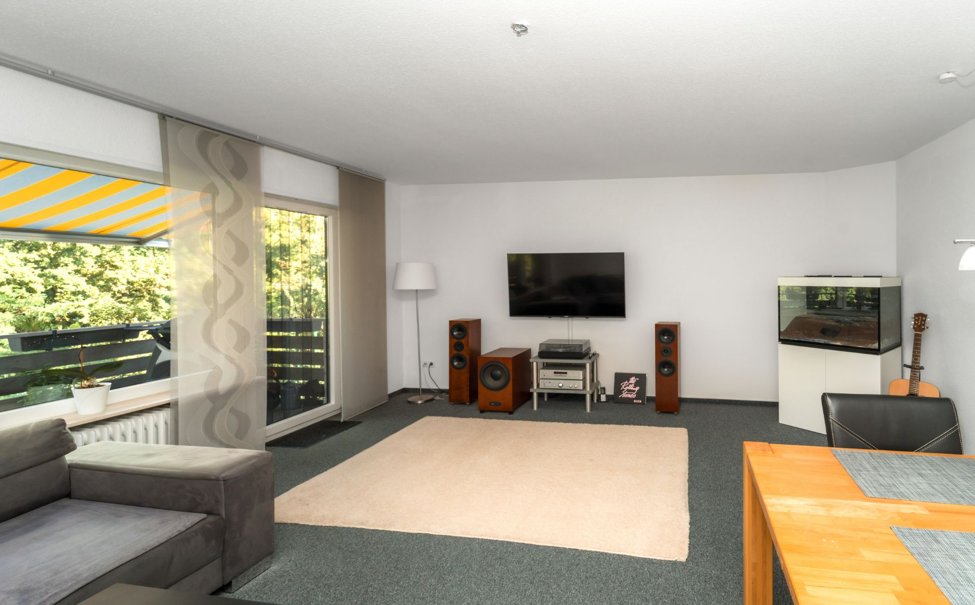 Wohnzimmer mit grauem Sofa, Teppich, Fernseher, Lautsprechern, Aquarium und großem Fenster.