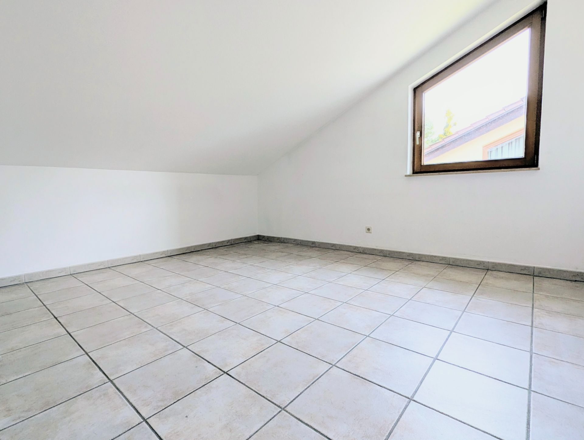 Leerer Raum mit weißer Wand, gefliestem Boden und schräger Decke, Fenster an der rechten Wand.