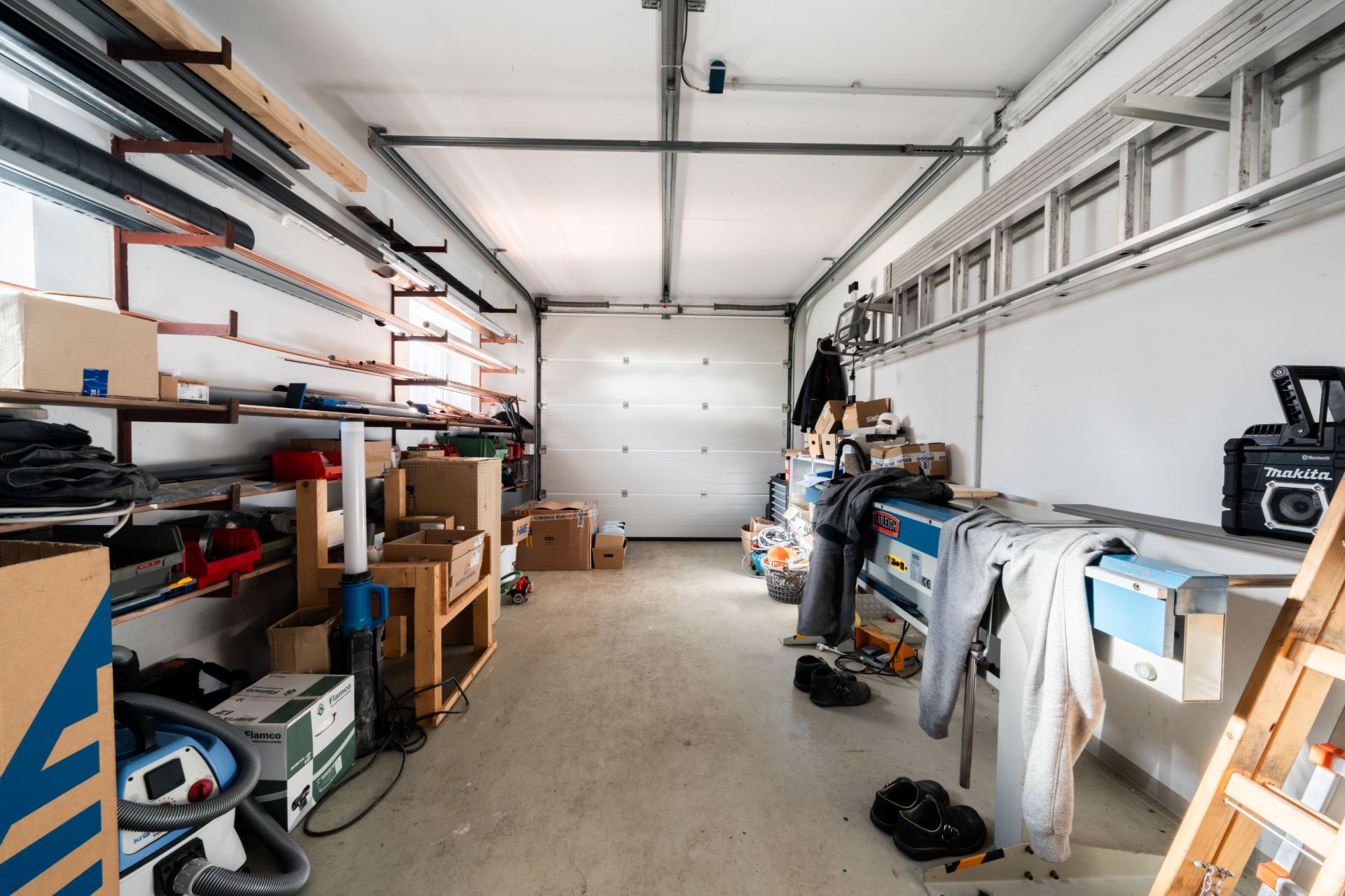 Garage mit Regalen, Werkzeugen, Kartons und einer Leiter an der Wand.