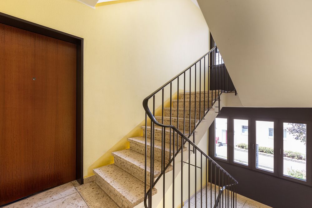 Treppenhaus mit brauner Wohnungstür, gelben Wänden und Fenstern, die nach draußen führen.