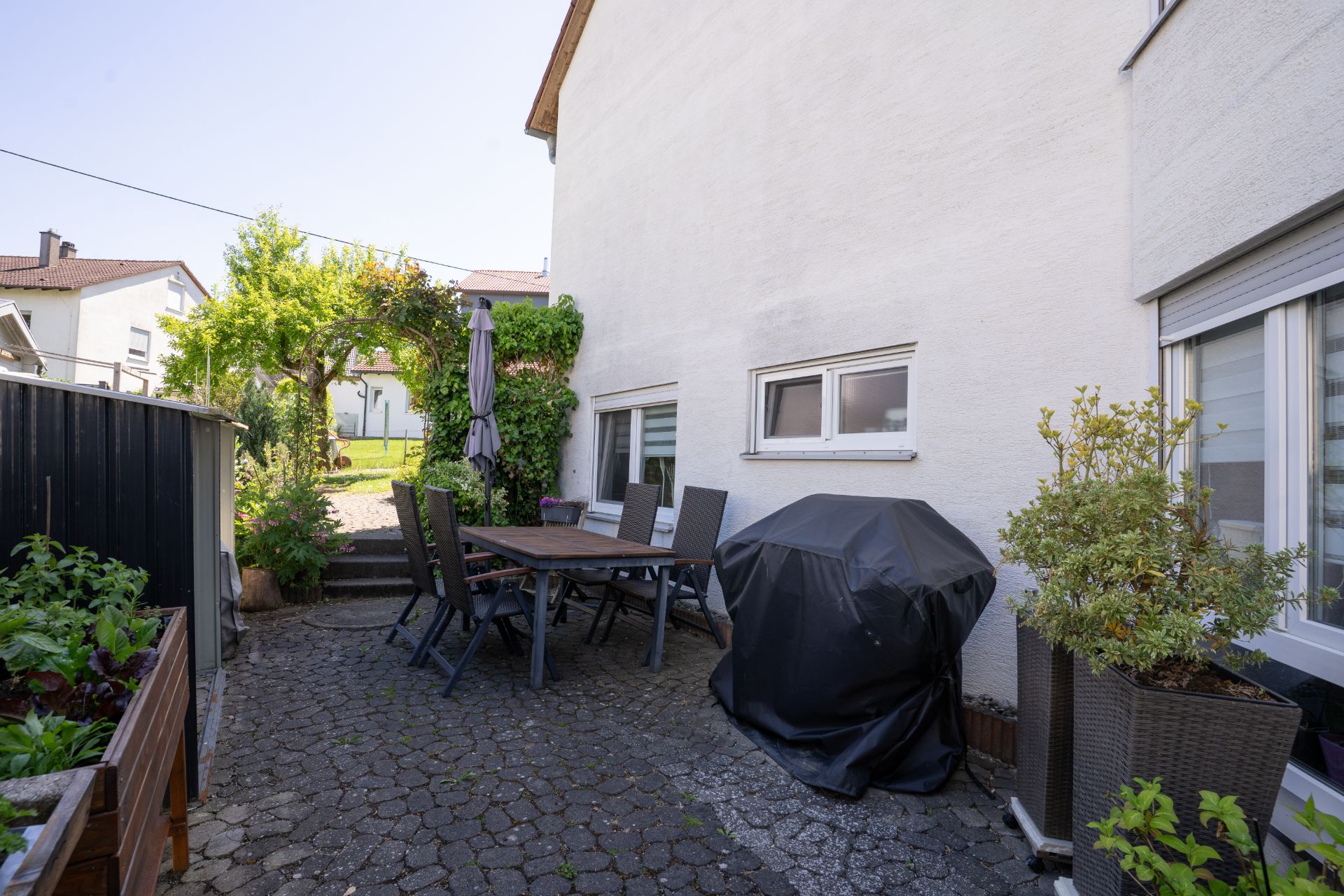 Gepflasterte Terrasse mit Tisch, Stühlen, Grill und Pflanzen neben einem Wohnhaus.