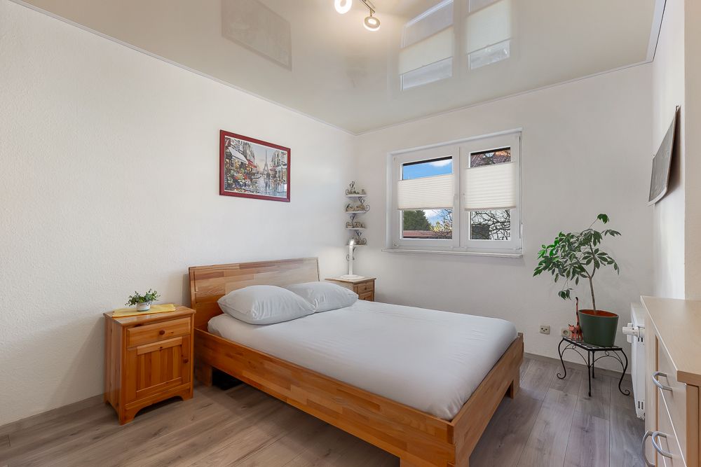 Helles Schlafzimmer mit Doppelbett, Holzmöbeln, Pflanze und großem Fenster.